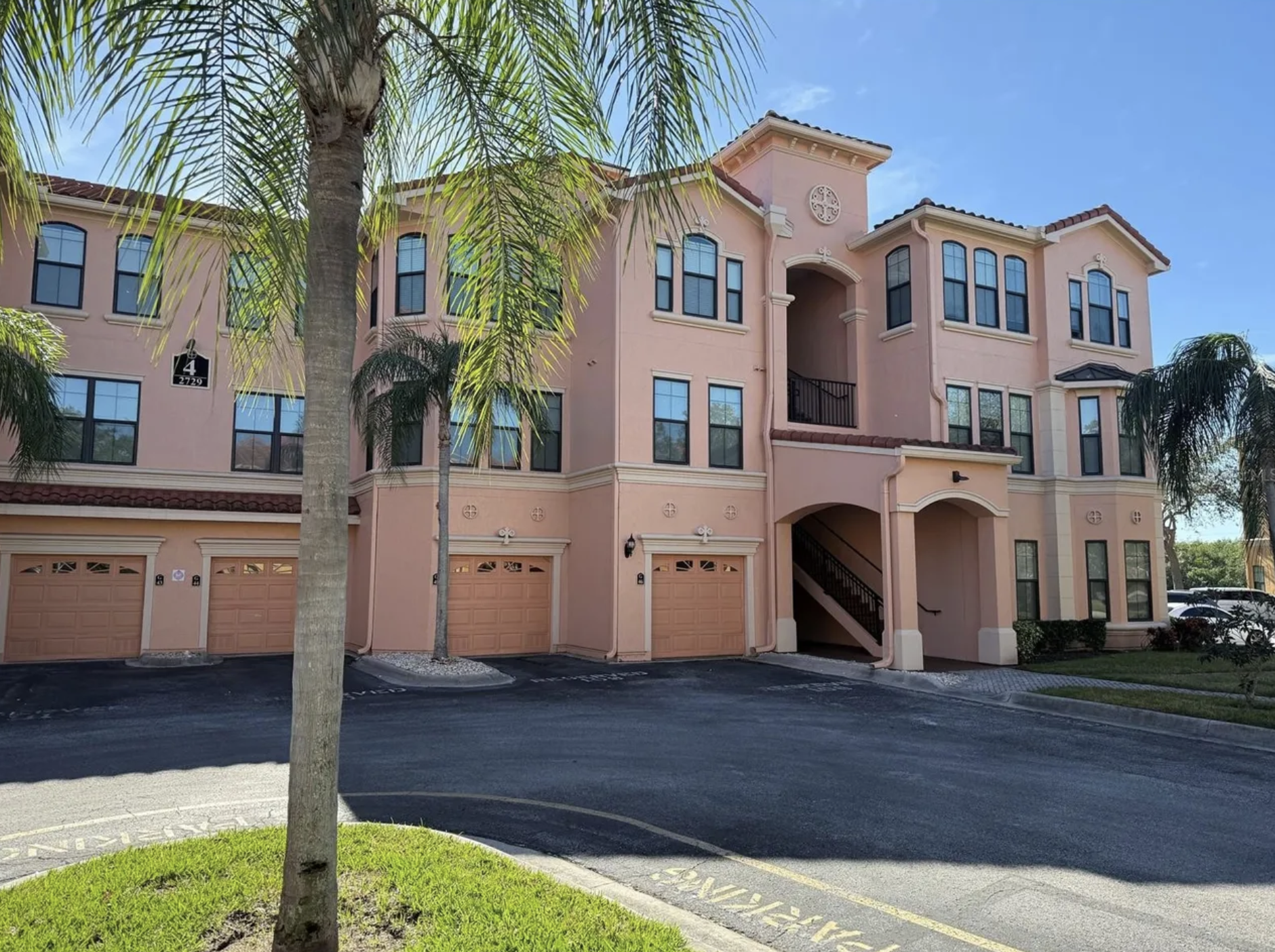 A three-story pink apartment building with multiple black-framed windows and a staircase entrance, surrounded by palm trees and a parking lot with marked spaces.