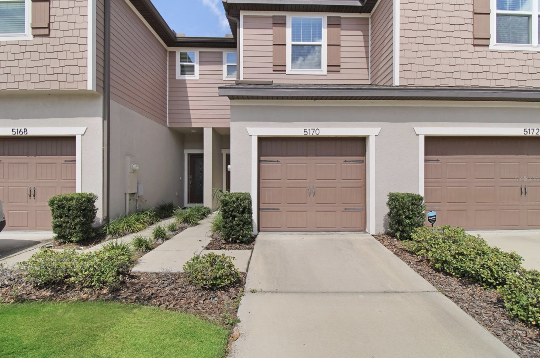 Front view of a modern residential townhouse with brown garage doors, pink siding, and a small front yard with bushes and a sidewalk.