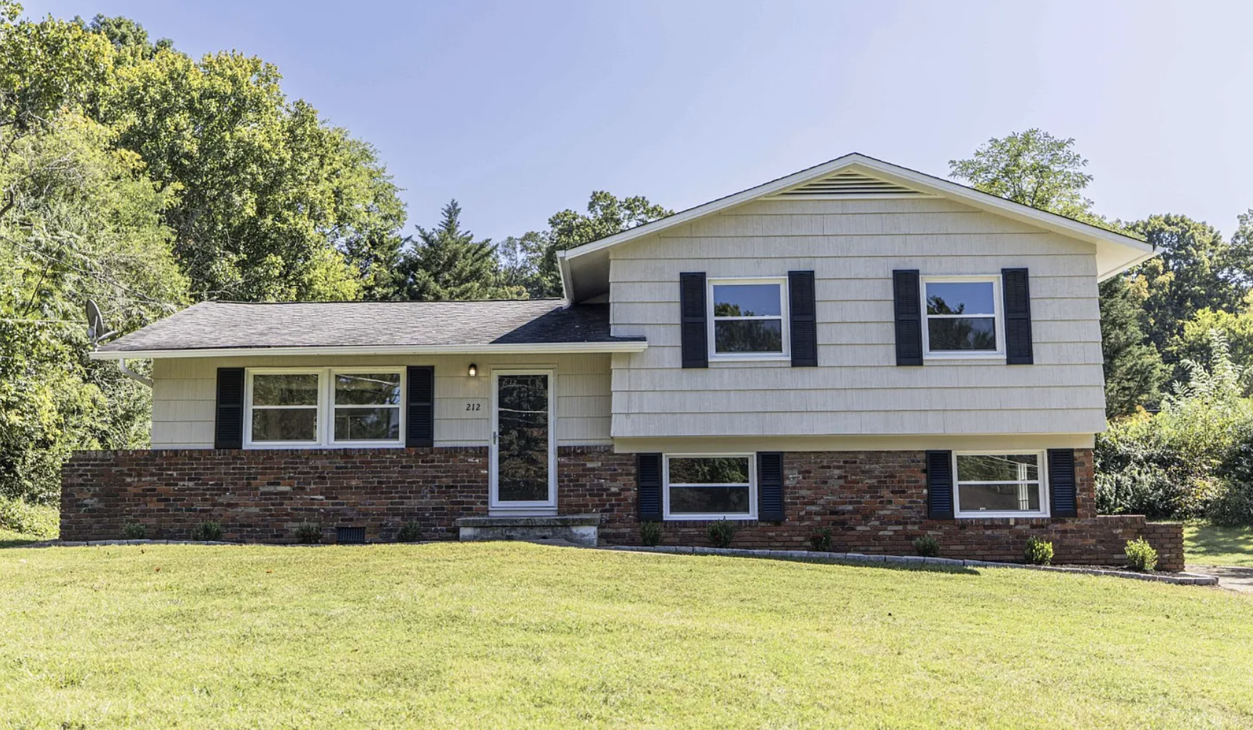 Front of a two-story house with beige siding, black shutters, and brick lower level. There is a lawn in front, trees behind, and a clear blue sky.