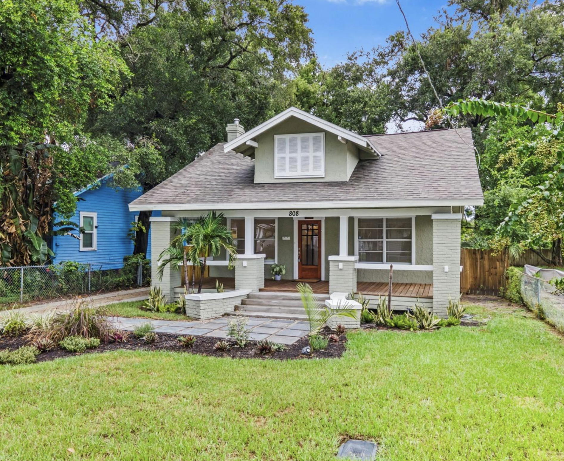Front view of a light green house with a gabled roof, a small porch with stairs, and surrounded by a lawn and garden with various plants.