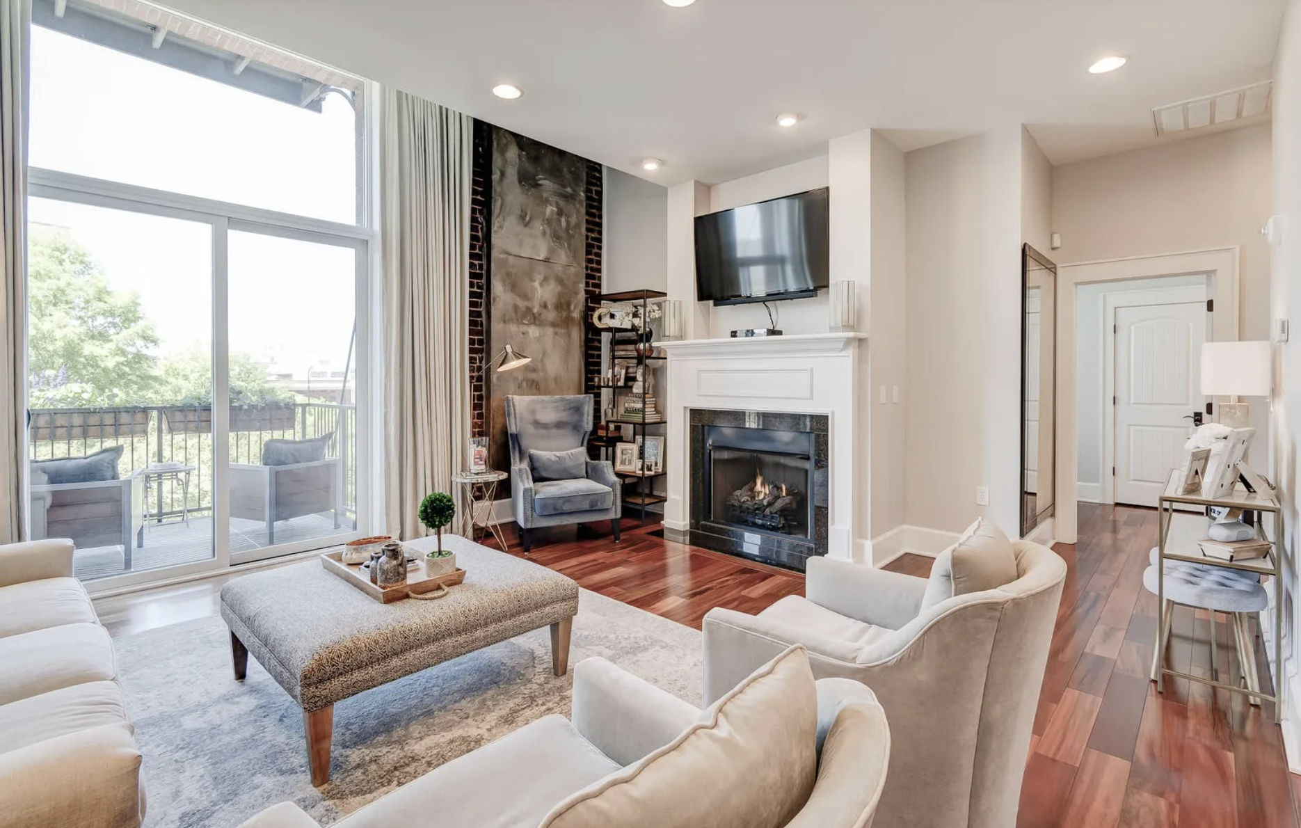 Living room with large sliding glass door leading to a balcony, beige sofas, a gray armchair, a fireplace, a TV mounted above the fireplace, a wooden shelf with books, and a small side table with a lamp. There are hardwood floors and a light-colored area rug.