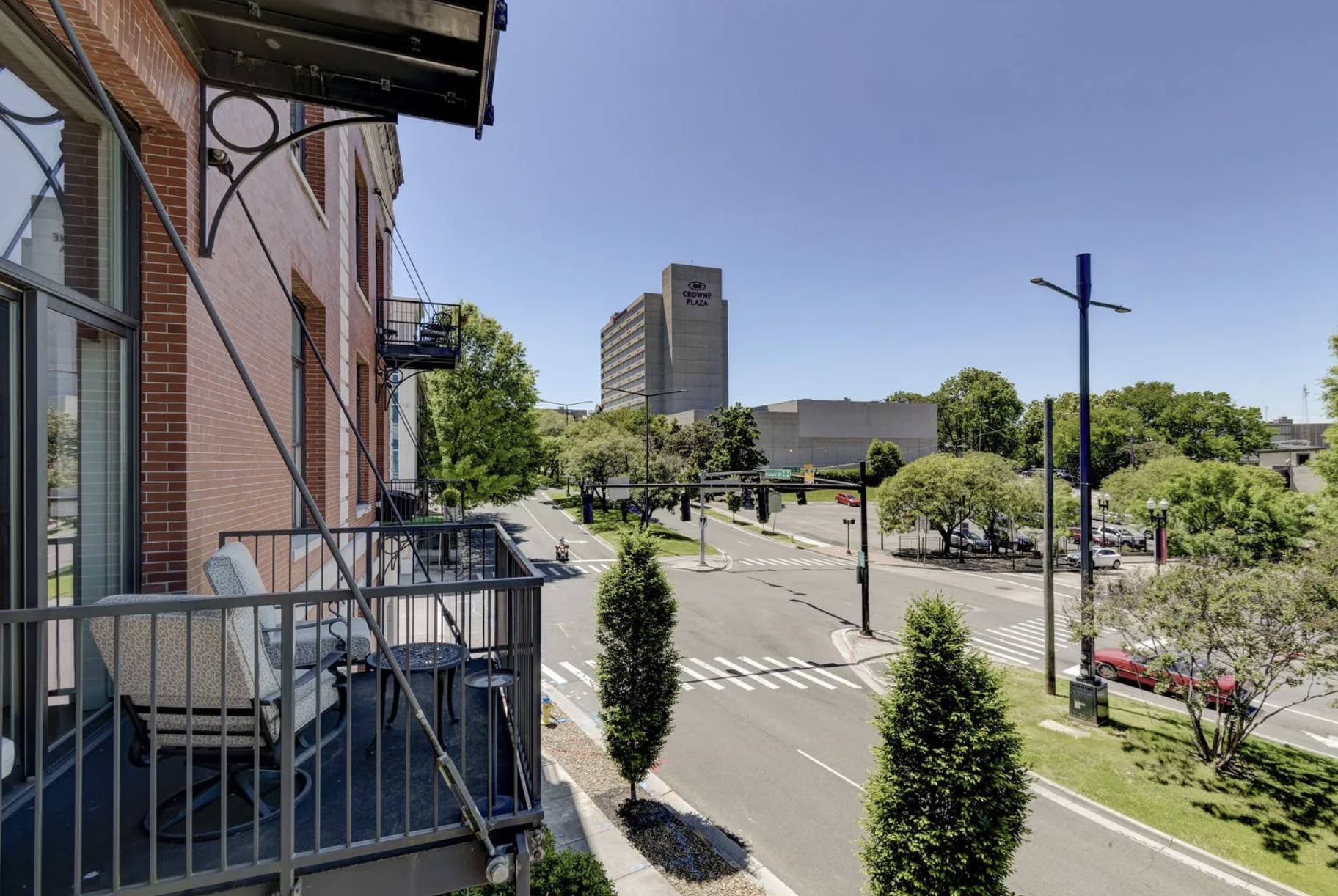 City street view from a balcony in a residential area with trees, cars, and tall buildings, including Crowne Plaza hotel in the background on a sunny day.