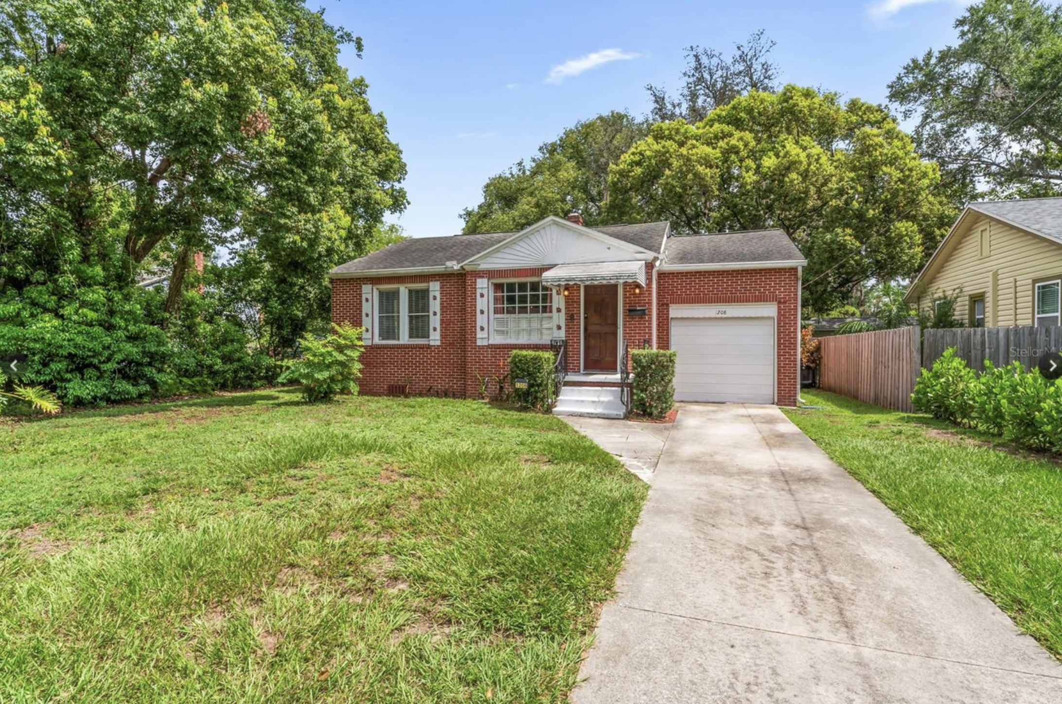 A single-story brick house with a white garage door, front yard with green grass, large trees in the background, and a concrete walkway leading to the front door.