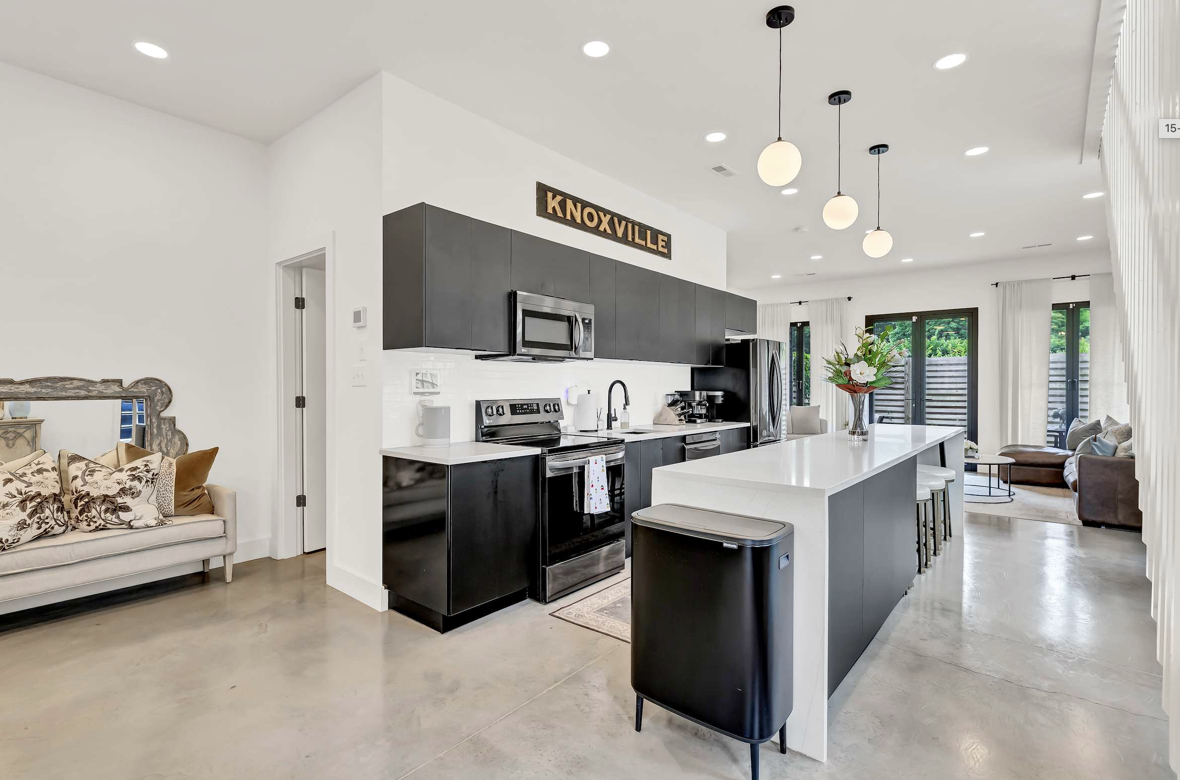 Open-concept modern kitchen with black cabinets, stainless steel appliances, and white island. Adjacent living area with a cream sofa and decorative pillows, a large bouquet of flowers on the table, and glass doors leading outside with lush greenery.