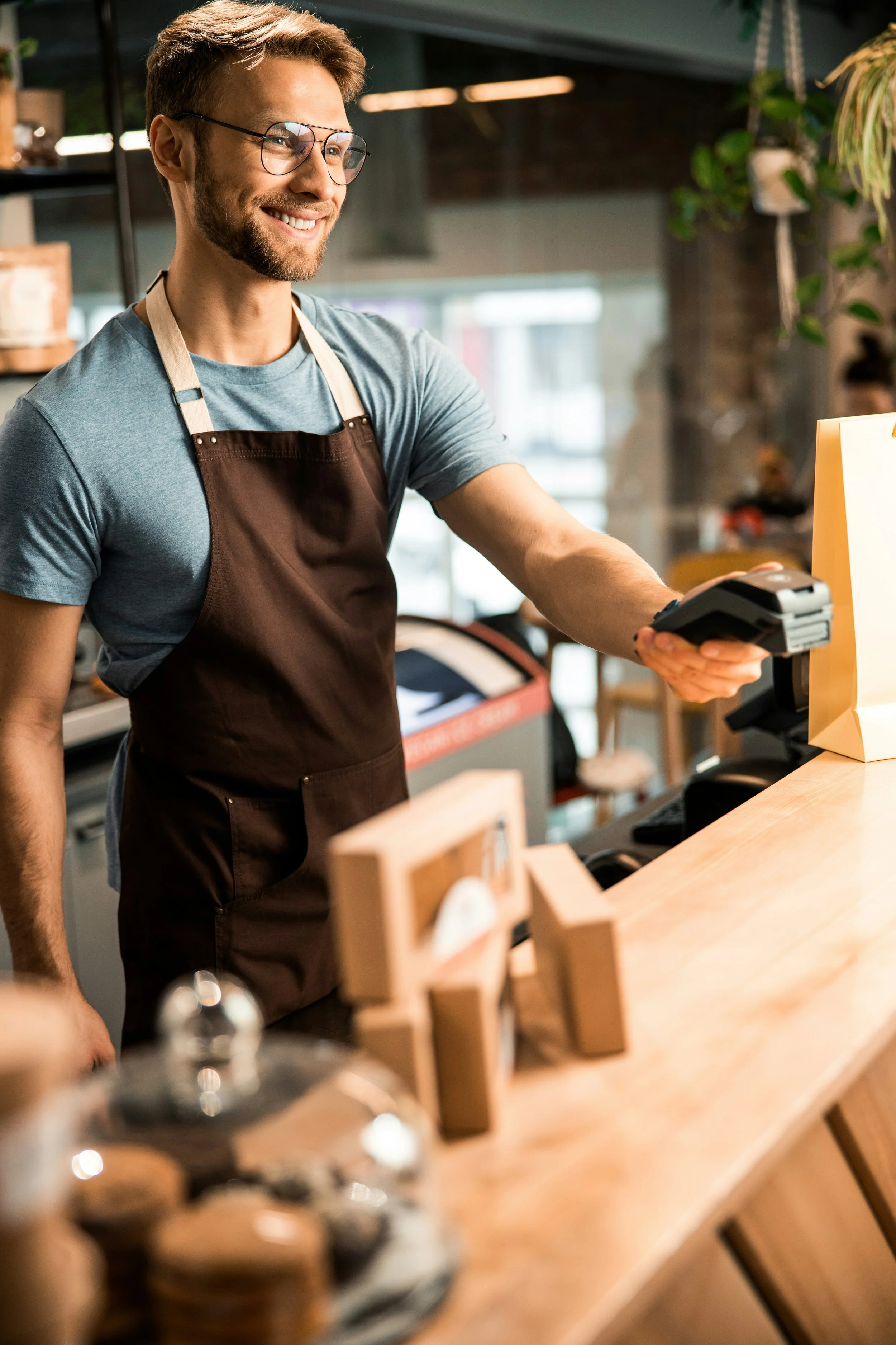 A smiling man with glasses and a beard working at a counter in a coffee shop or cafe, wearing a brown apron over a blue t-shirt.