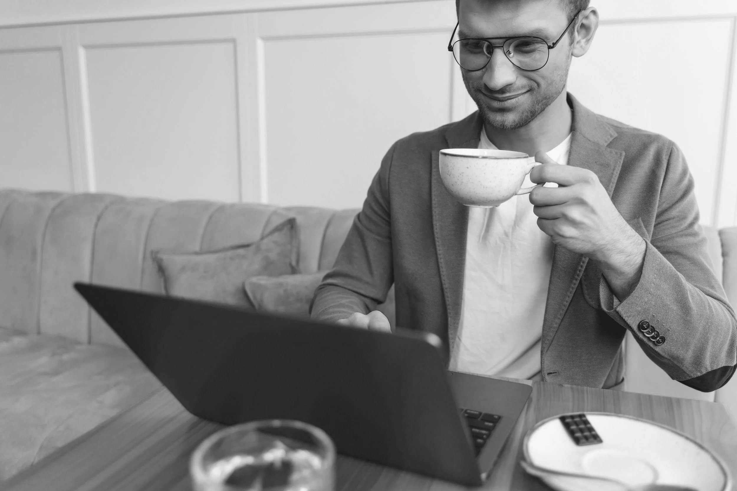 A man with glasses and a beard sitting at a table, holding a cup and working on a laptop.