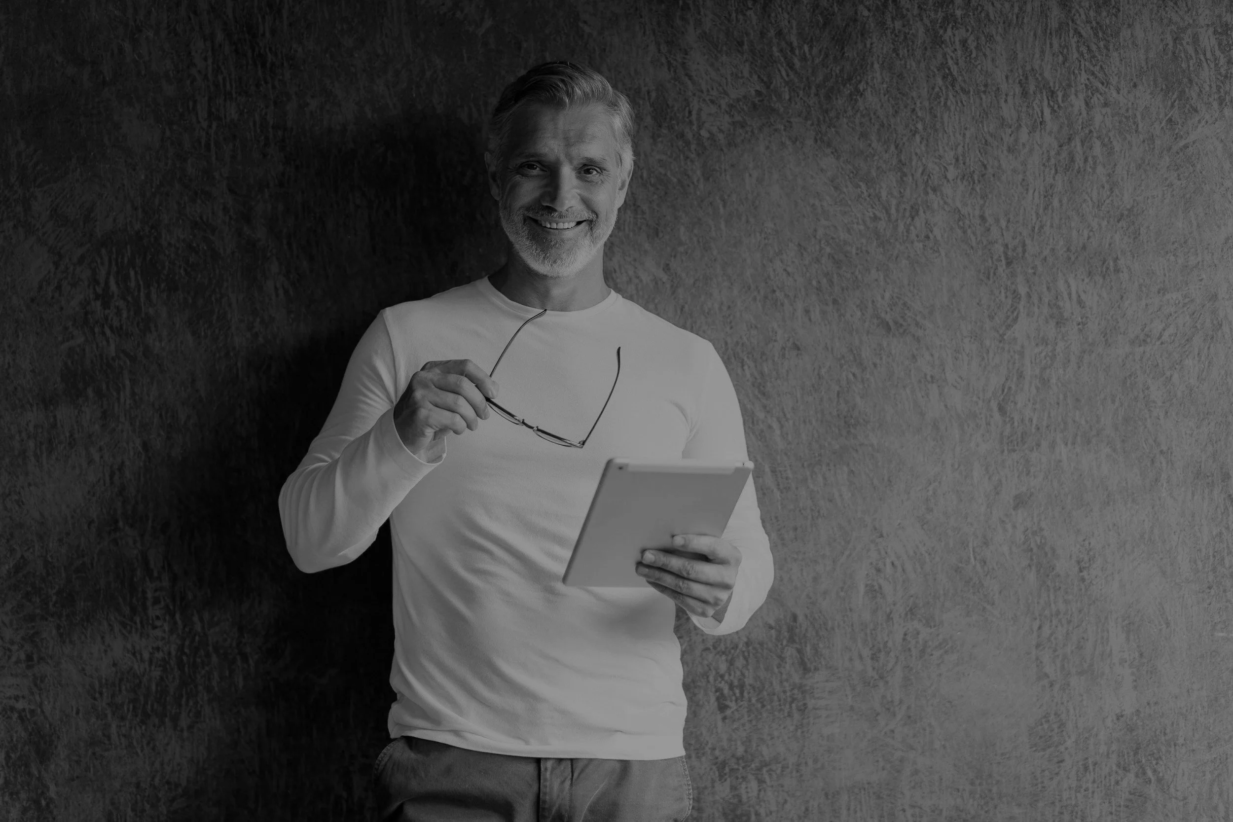 A smiling older man with gray hair and a beard holding glasses and a tablet, standing against a textured wall.