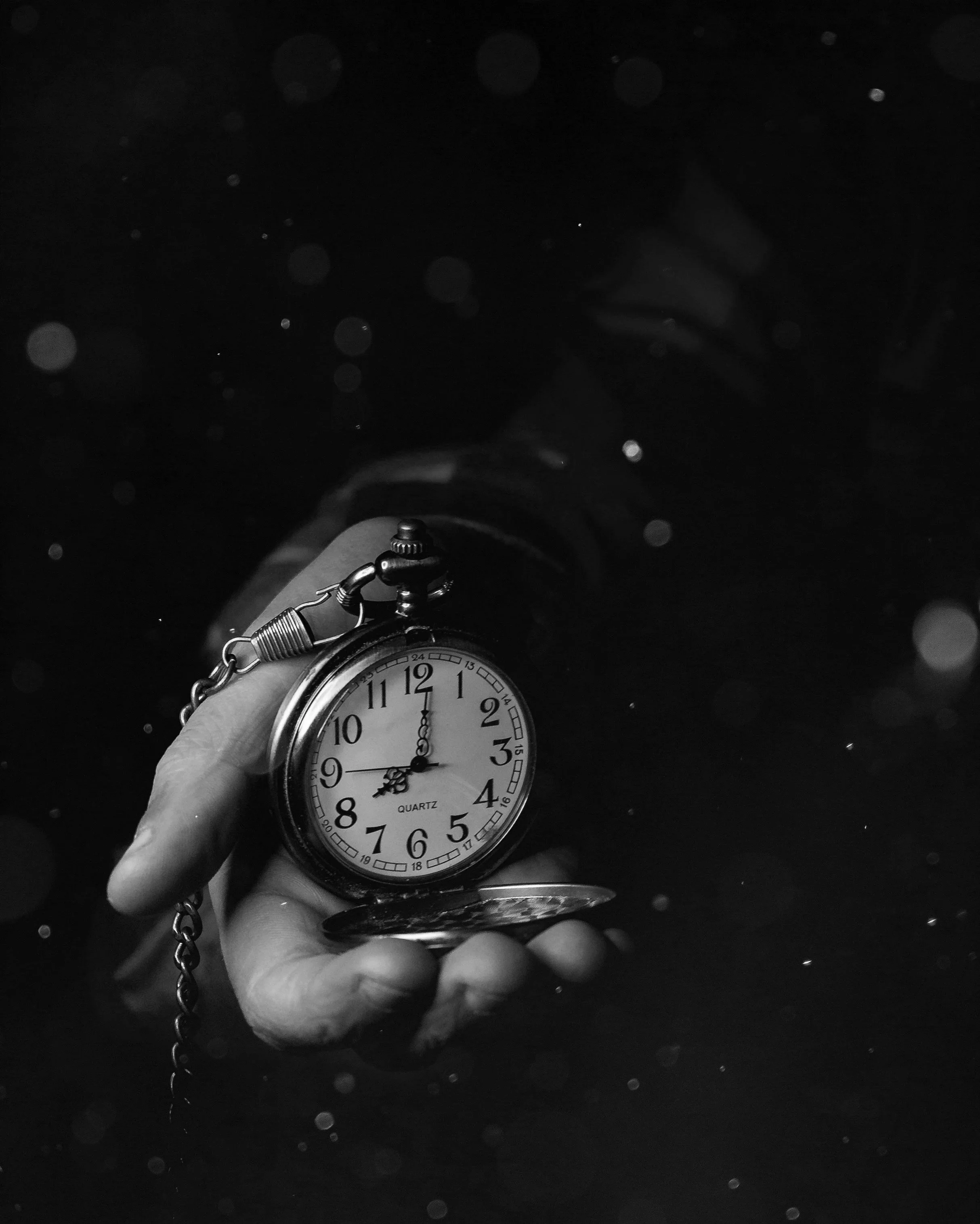 A person holding a vintage pocket watch with a chain, showing the time as 11:02, in a dark background with floating light particles.