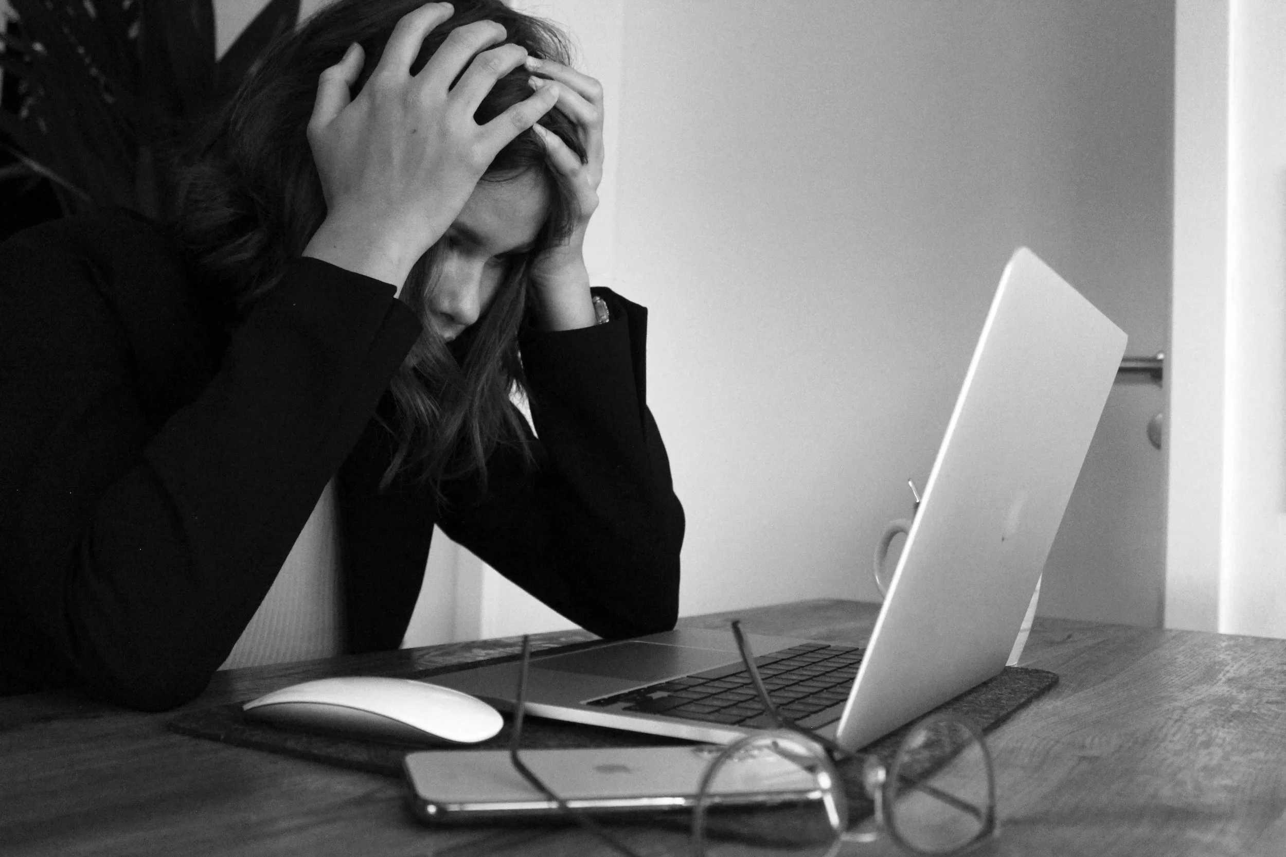 Frustrated woman with hands on her head sitting at a table with a laptop, smartphone, and glasses nearby in black and white.