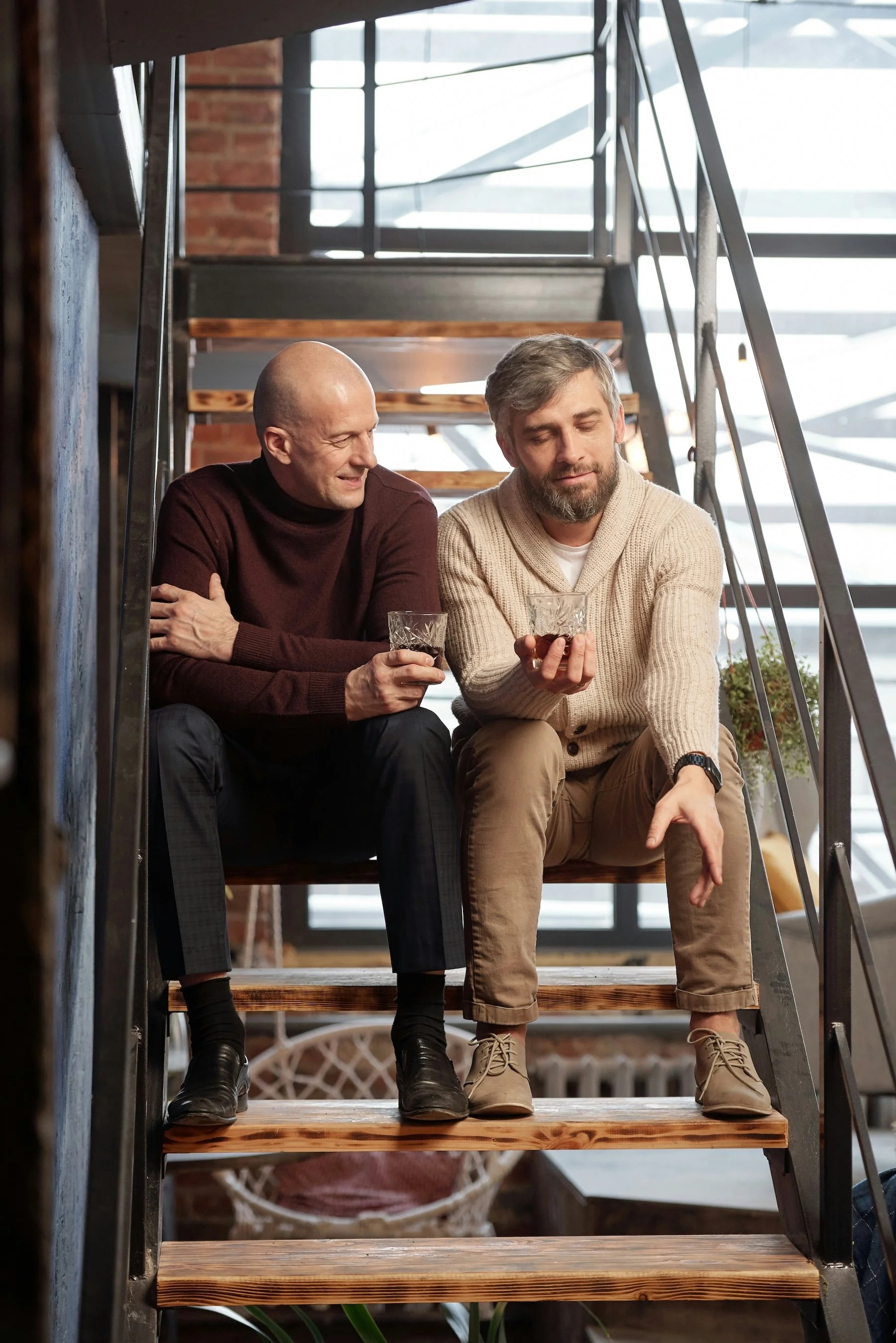 Two men sitting on stairs inside a modern building with large windows, holding glasses of a dark beverage, engaged in a conversation.