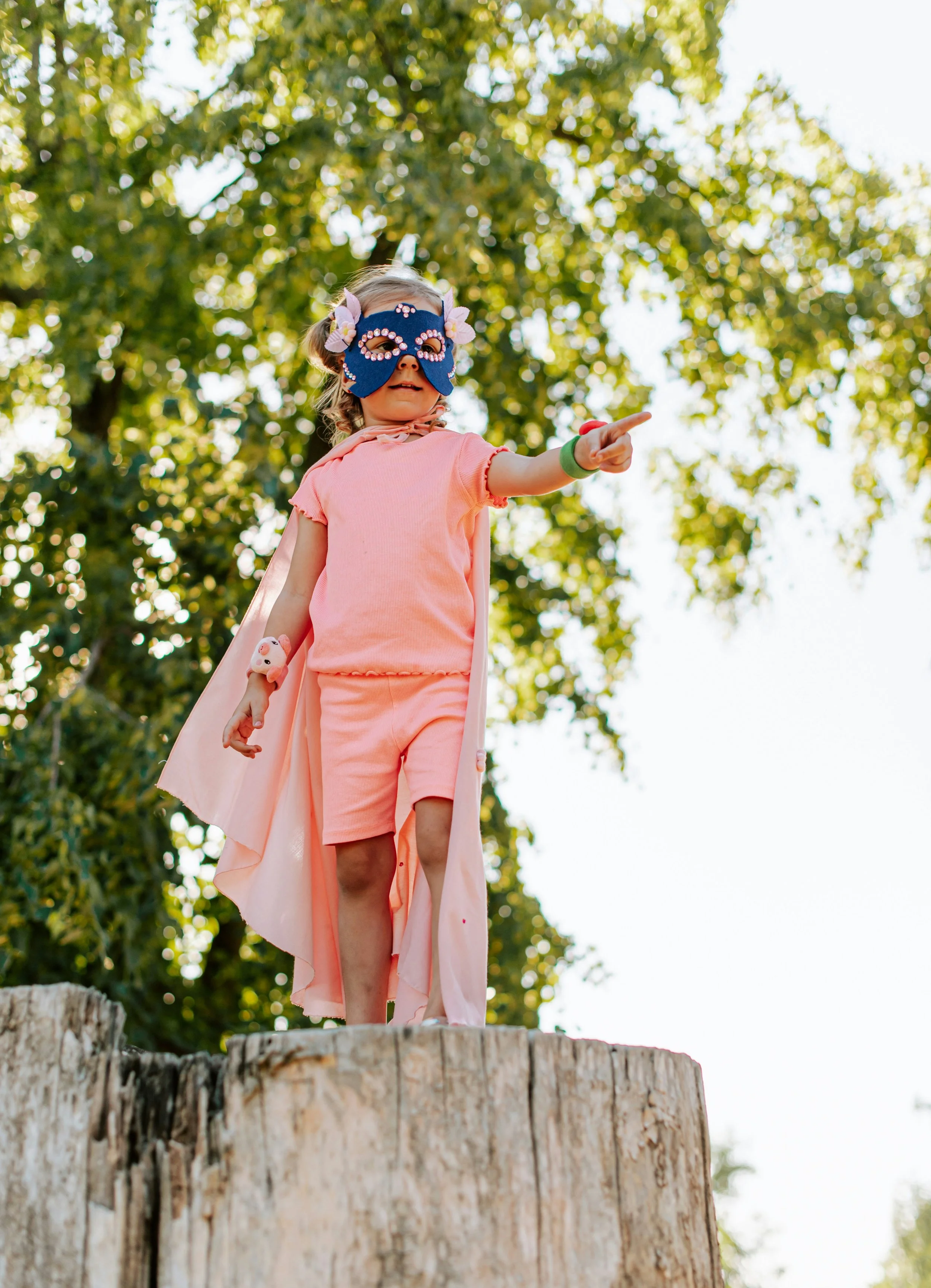 A young girl wearing a blue butterfly mask, pink cape, and matching pink outfit, standing on top of a large tree stump outdoors, pointing at something in the distance, with green trees and sunlight in the background.