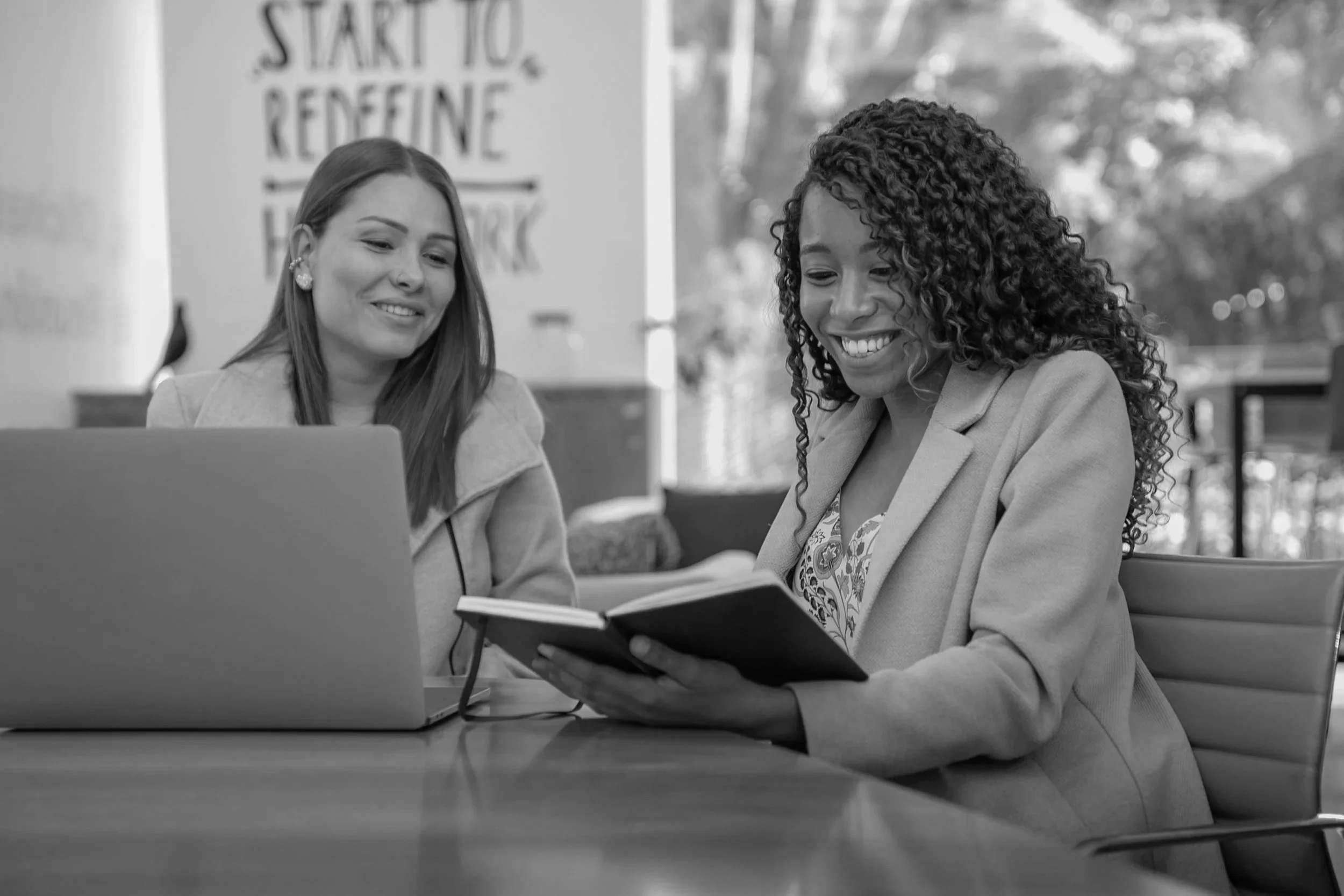 Two women sitting at a table, smiling and looking at a notebook, with a laptop open in front of them, in a bright indoor space.