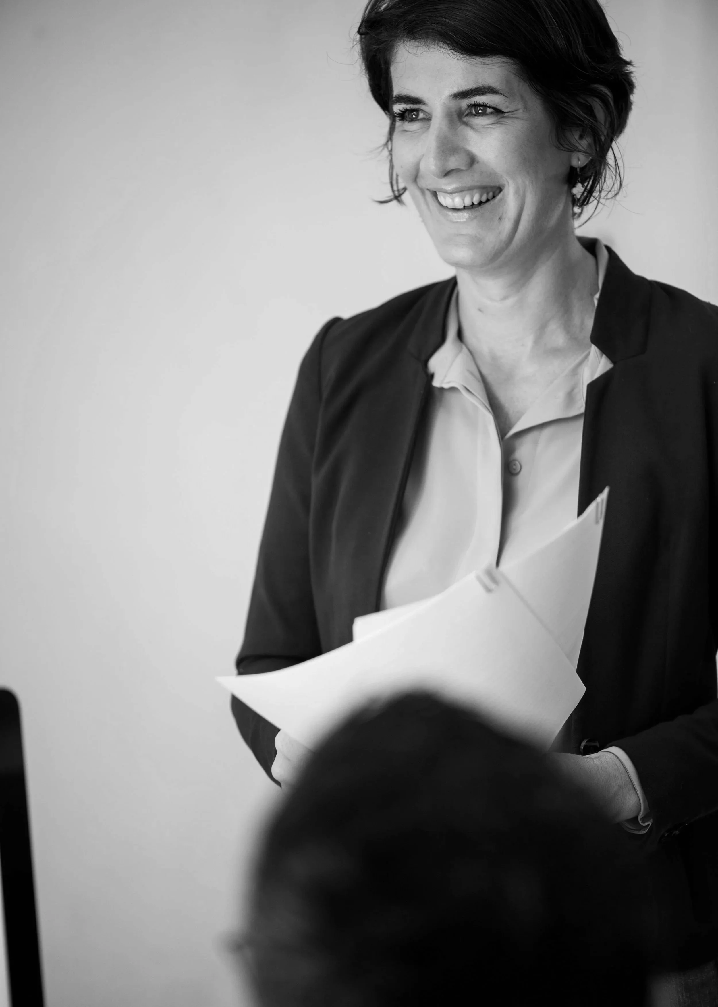 A woman with short dark hair smiling and holding papers, dressed in a dark blazer and light shirt, standing indoors.