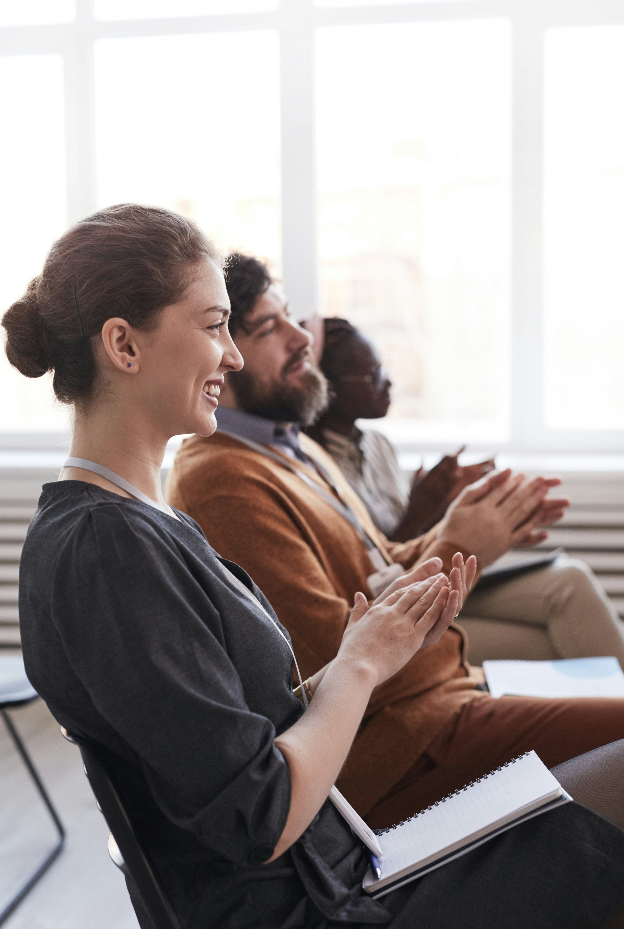 Three people sitting in a row, clapping, and smiling during a meeting or seminar in a bright room with large windows.