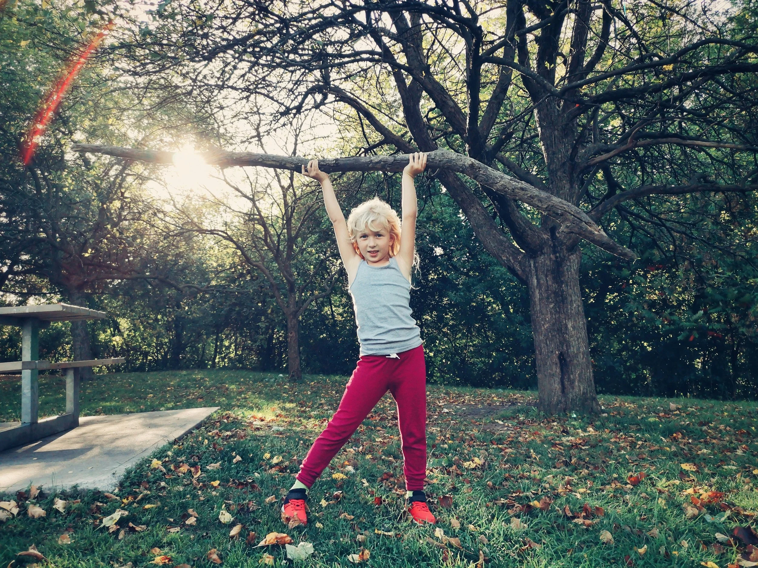 A young girl with blonde hair wearing a gray tank top and red pants stands outdoors in front of a large tree, holding a branch above her head with both hands. The sunlight is shining through the trees, creating a bright, natural setting with fallen leaves on the grass.