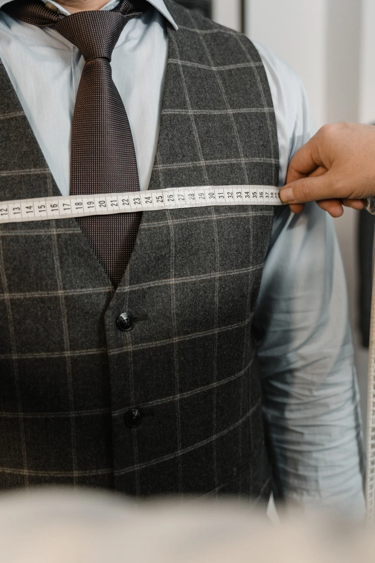 Person measuring chest with a tape measure, wearing a gray checkered vest and a dark tie over a white shirt.