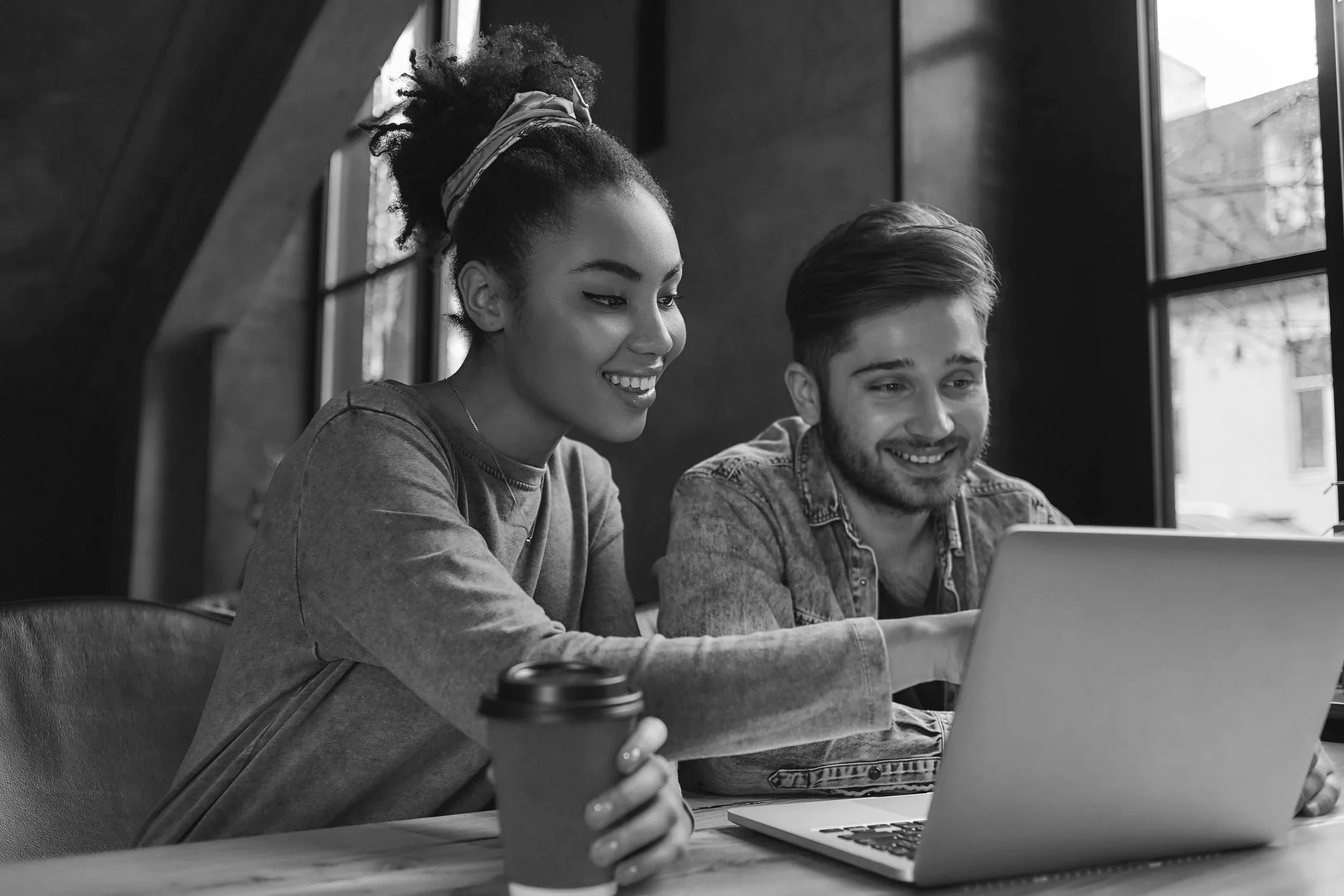 Two young adults, a woman and a man, sitting at a table and looking at a laptop. The woman is holding a coffee cup and smiling, while the man is also smiling. They are in a well-lit room with large windows.
