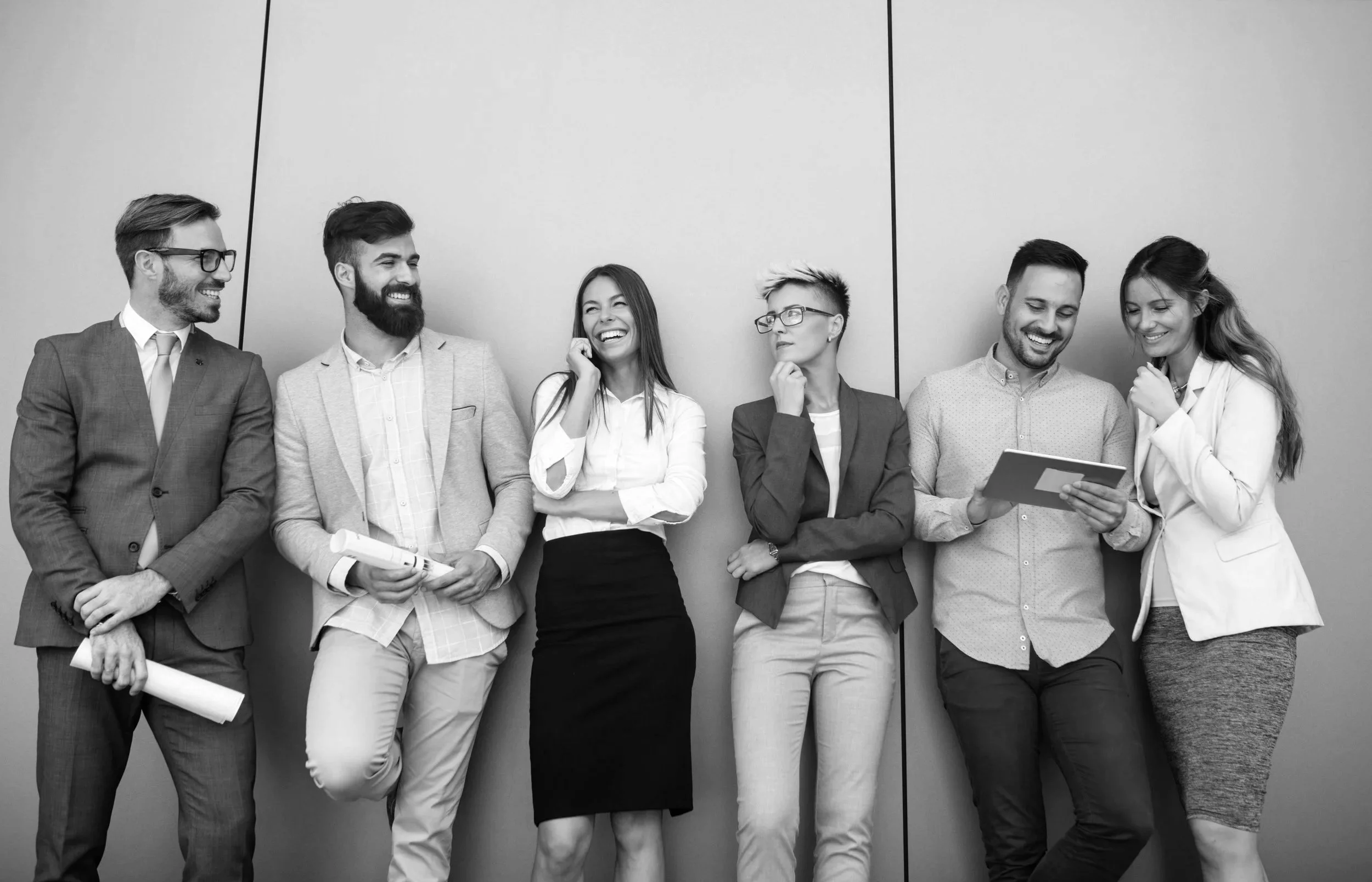 Six diverse adults in business attire standing and laughing together in front of a plain wall.