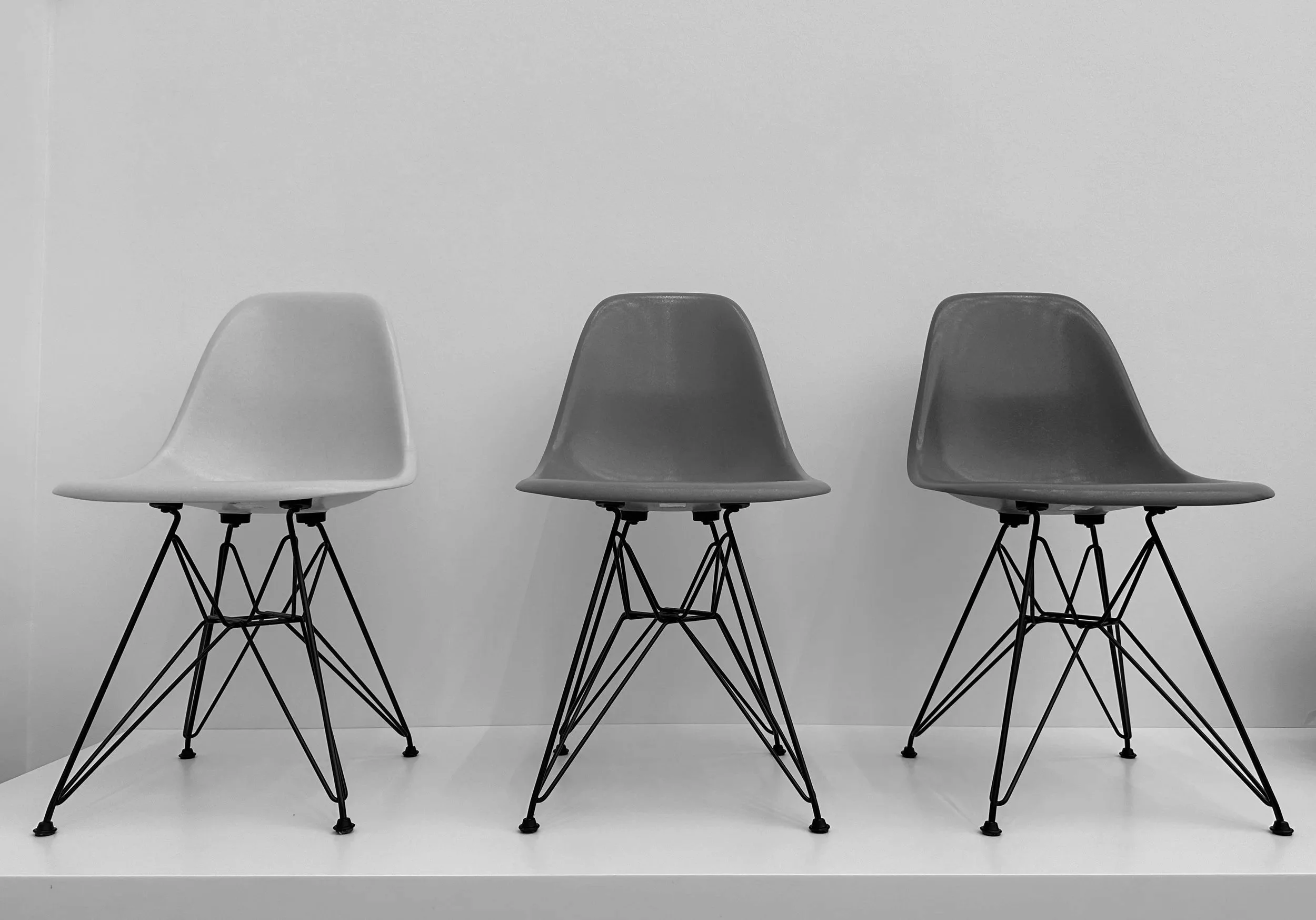 Three modern chairs with plastic seats on metal wire legs, arranged in a row on a white shelf against a plain background.