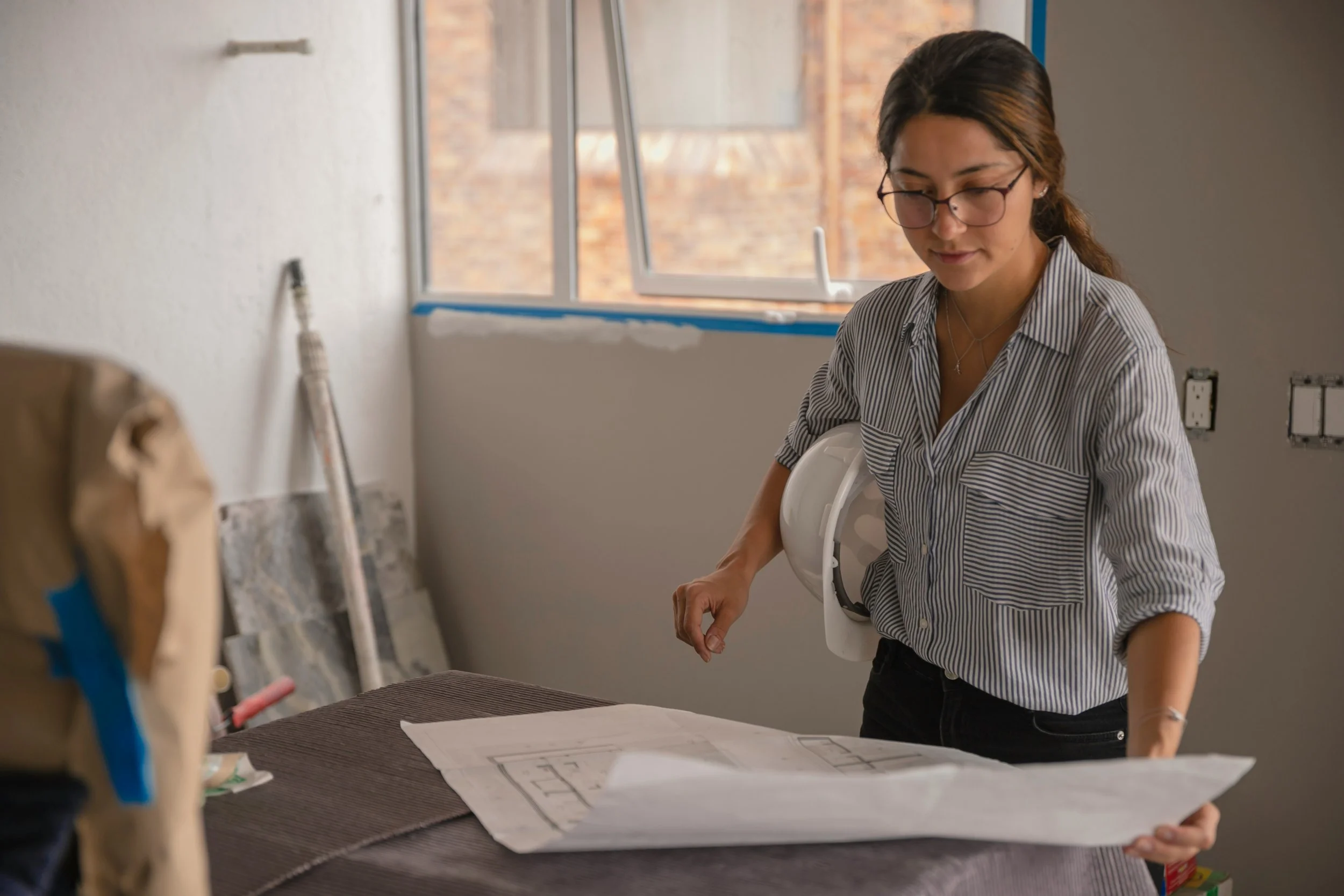A woman with glasses and a striped shirt looks down at blueprints or plans on a table in a room under construction, with construction materials and tools visible around her.