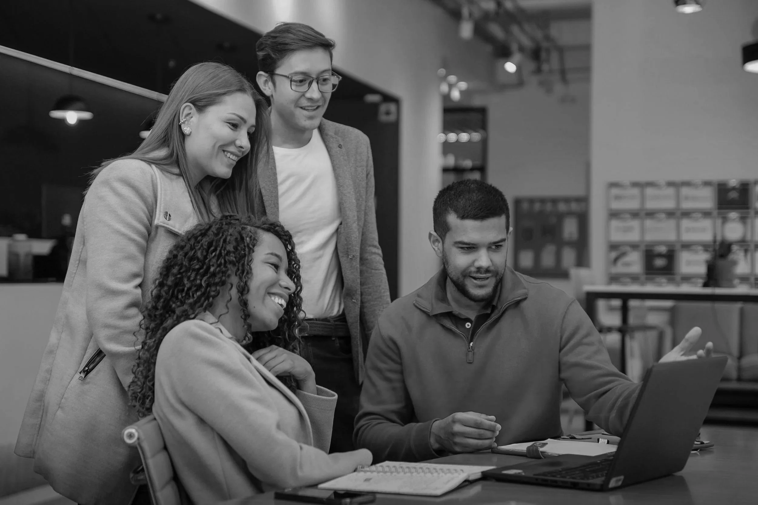 Four young adults gathered around a table, looking at a laptop screen, smiling and engaging with each other in a modern workspace.