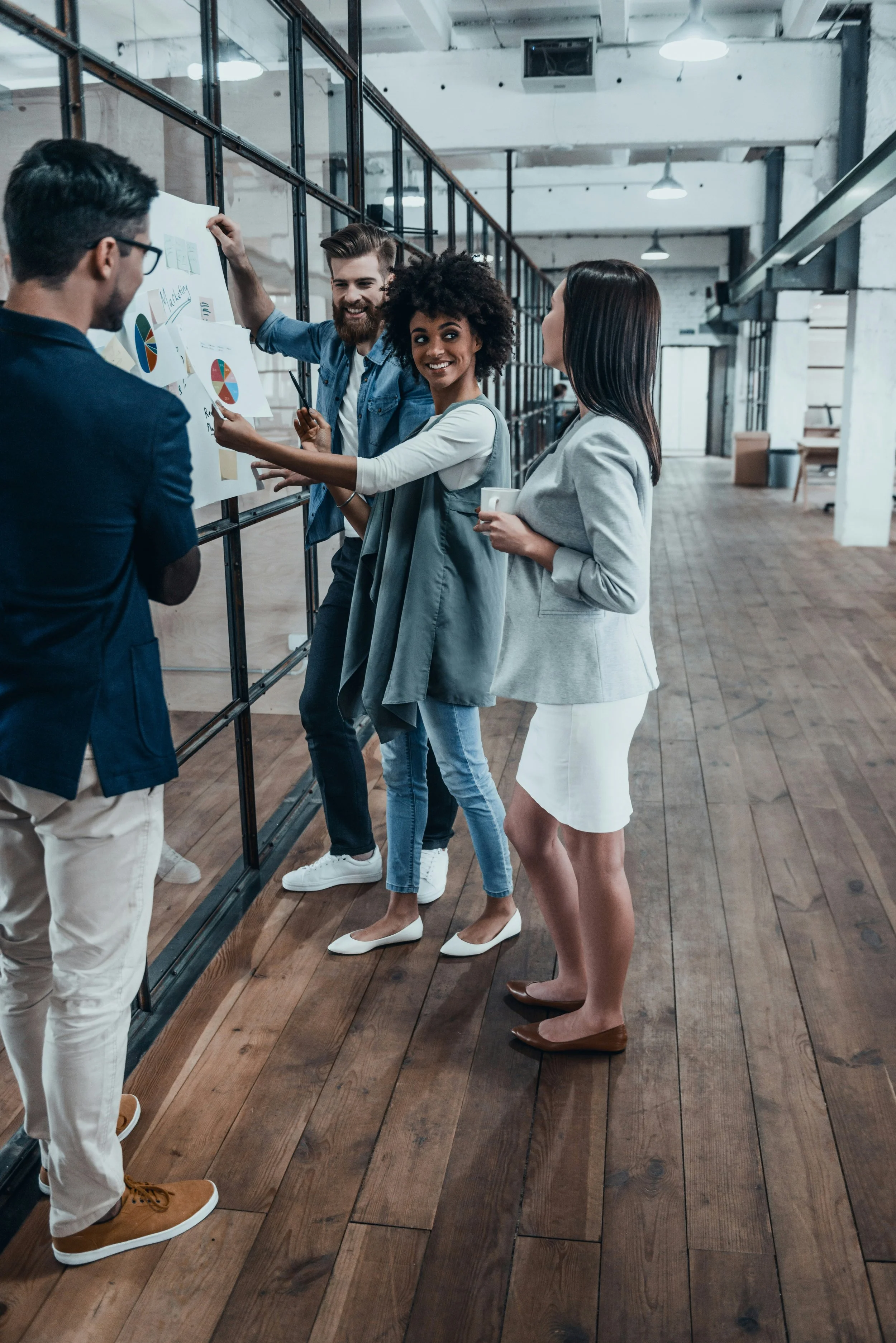 Four diverse young professionals in stylish office attire standing and discussing charts on a whiteboard in a modern, industrial-style office.
