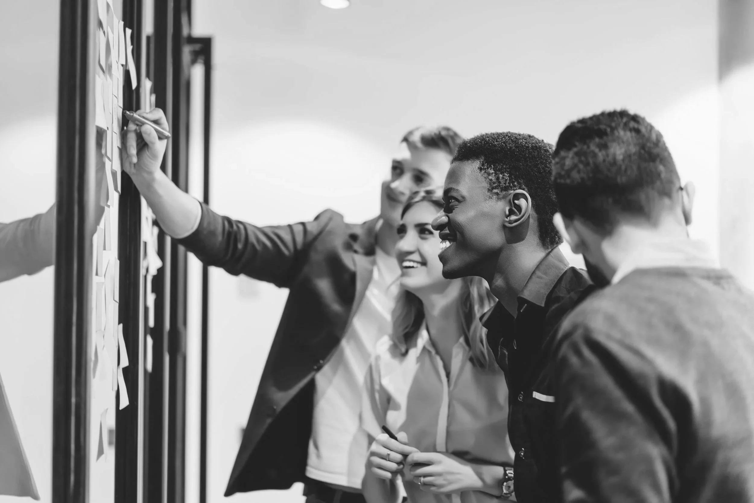 Four young professionals, two men and two women, are gathered around a board with sticky notes, discussing ideas and brainstorming.