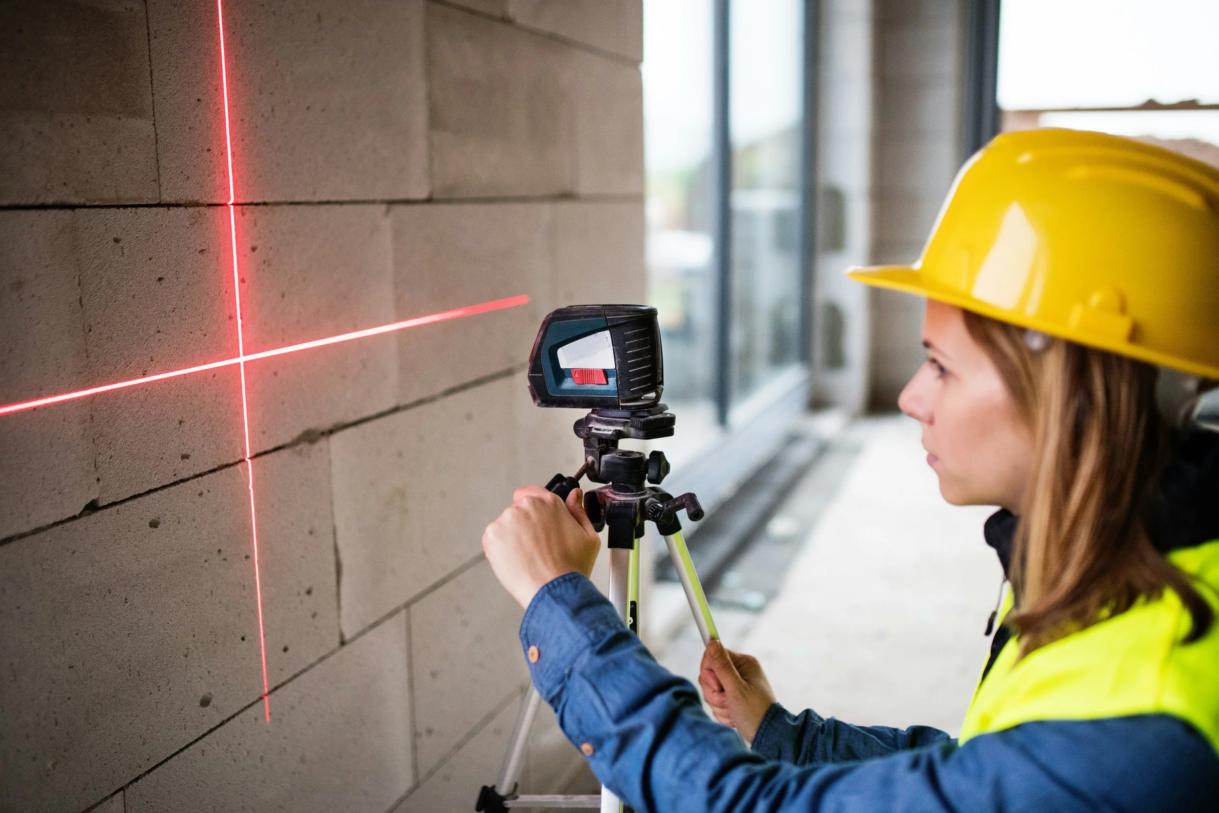 A woman in a yellow hard hat and a yellow safety vest is using a leveling tool to measure a brick wall with a red laser level projecting horizontal and vertical lines.