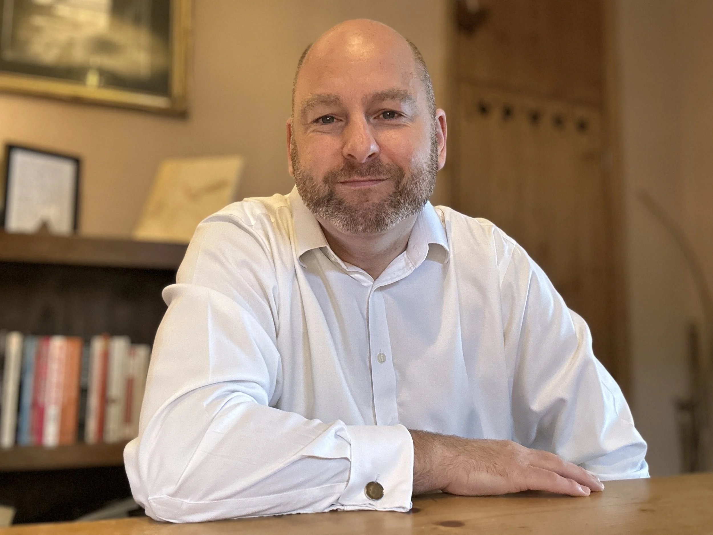 A man with a bald head and beard wearing a white shirt sitting at a wooden table in a room with bookshelves and framed pictures in the background.