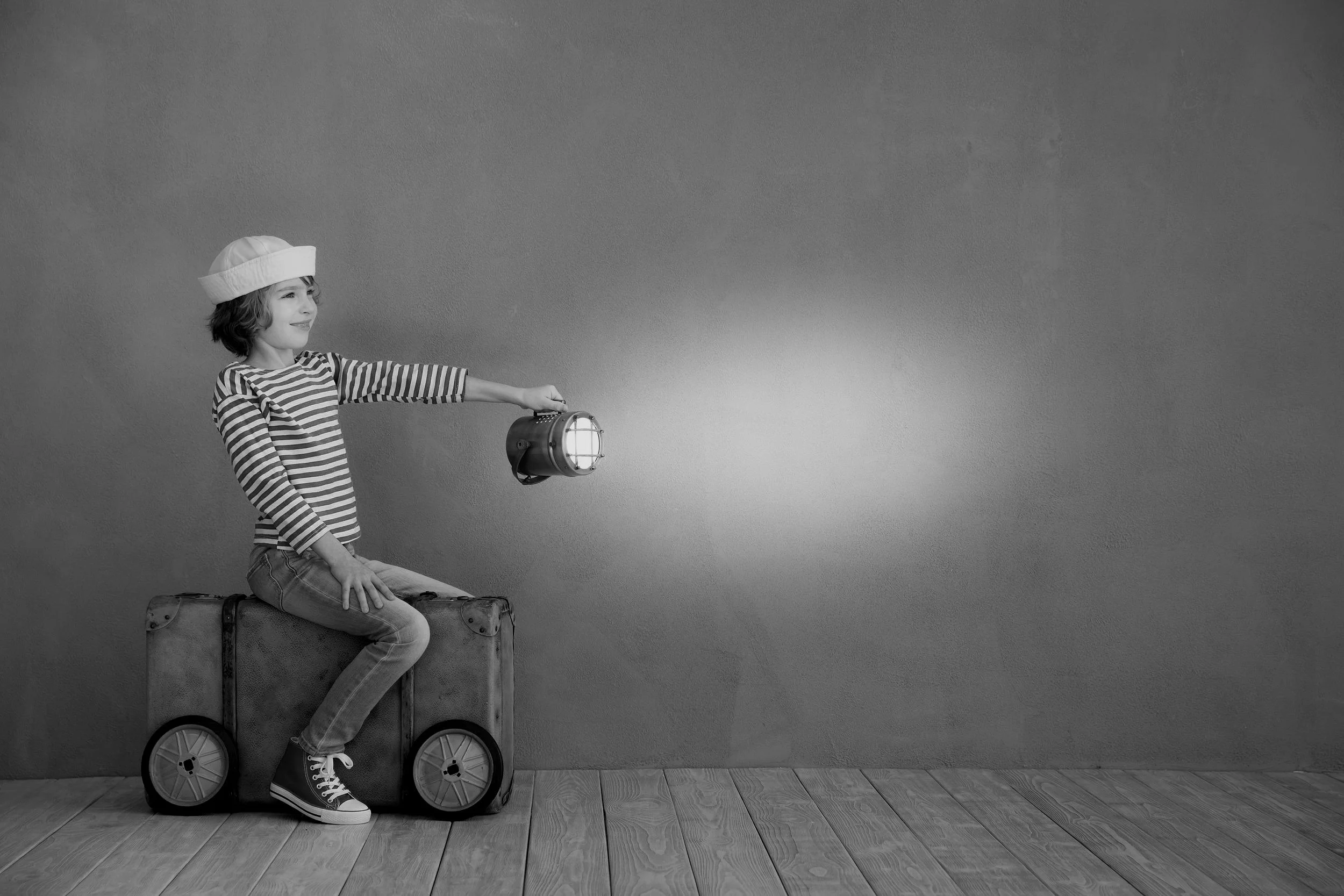 Child sitting on a suitcase, holding a flashlight, wearing a striped shirt and a hat, with a hardwood floor and plain wall background.