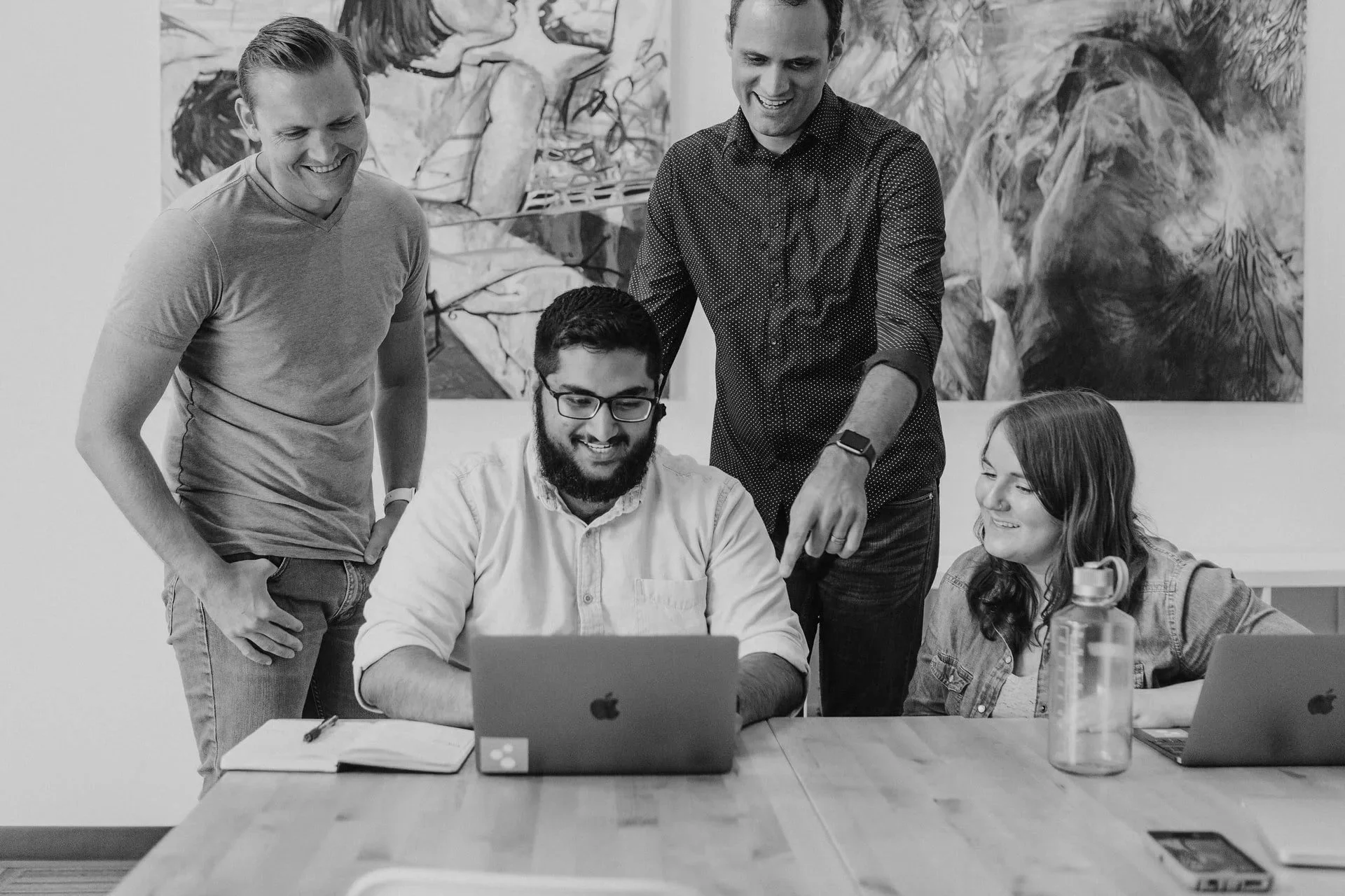 Four people in a meeting room, three standing and one sitting, looking at a laptop and smiling, with notebooks and water bottles on the table, and abstract art on the wall in the background.