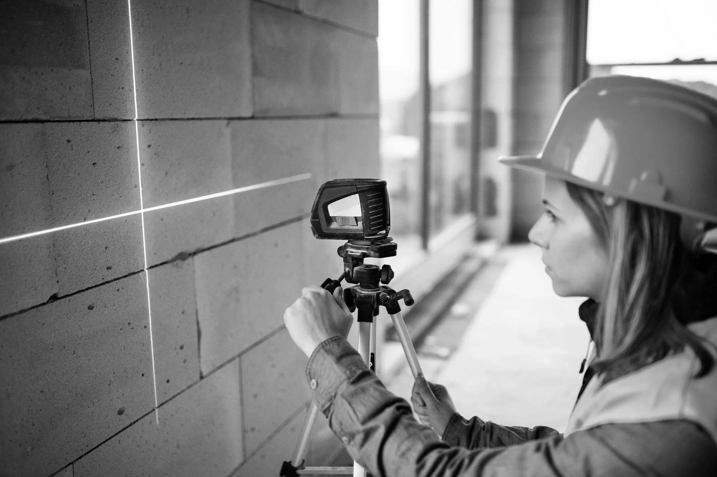 A woman wearing a construction helmet uses a laser level to measure on a wall at a construction site.