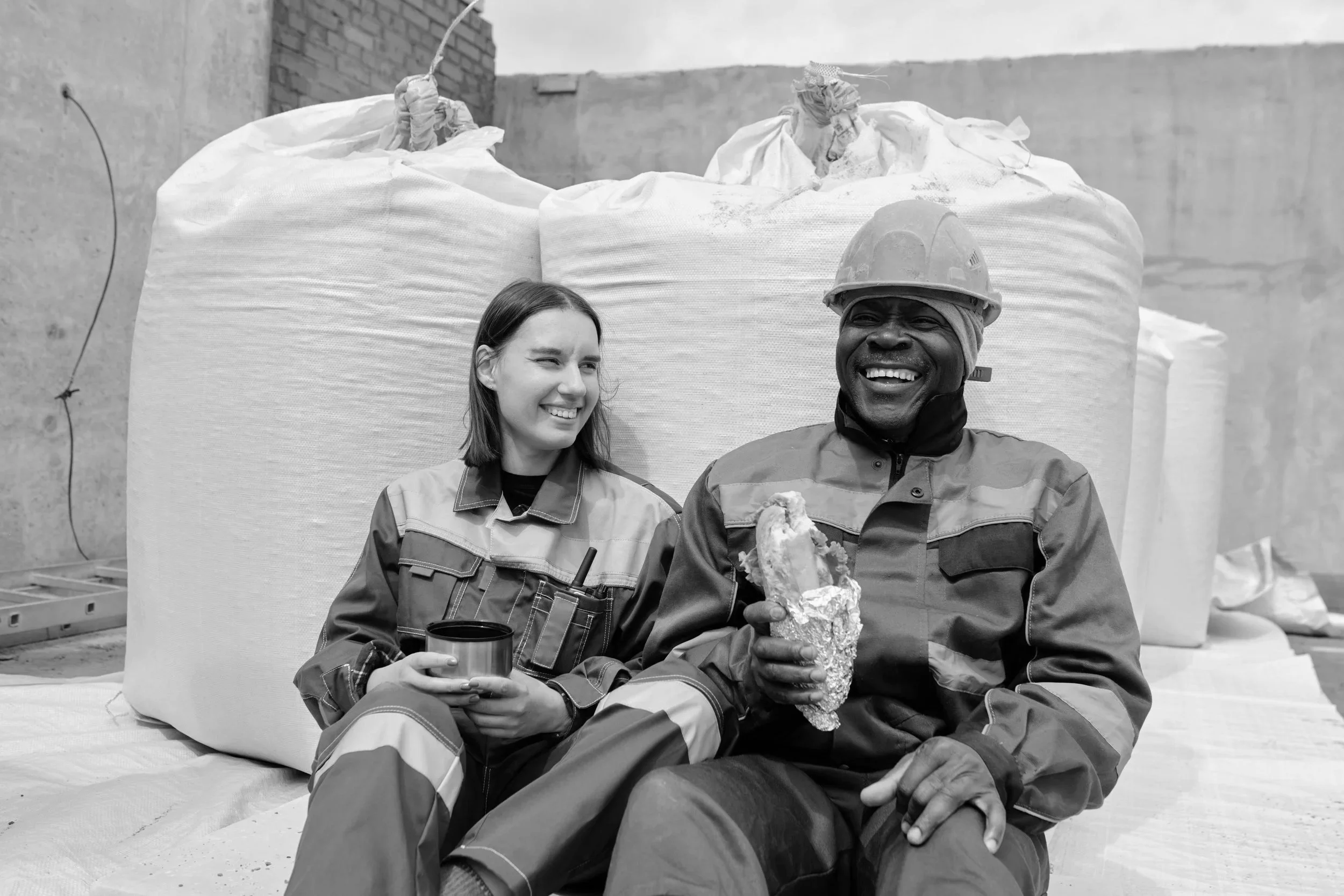 Two workers, a woman and a man, sitting on the ground, smiling. The man is holding a sandwich, and the woman is holding a cup. They are wearing work uniforms and safety gear, with large bags or bundles behind them.