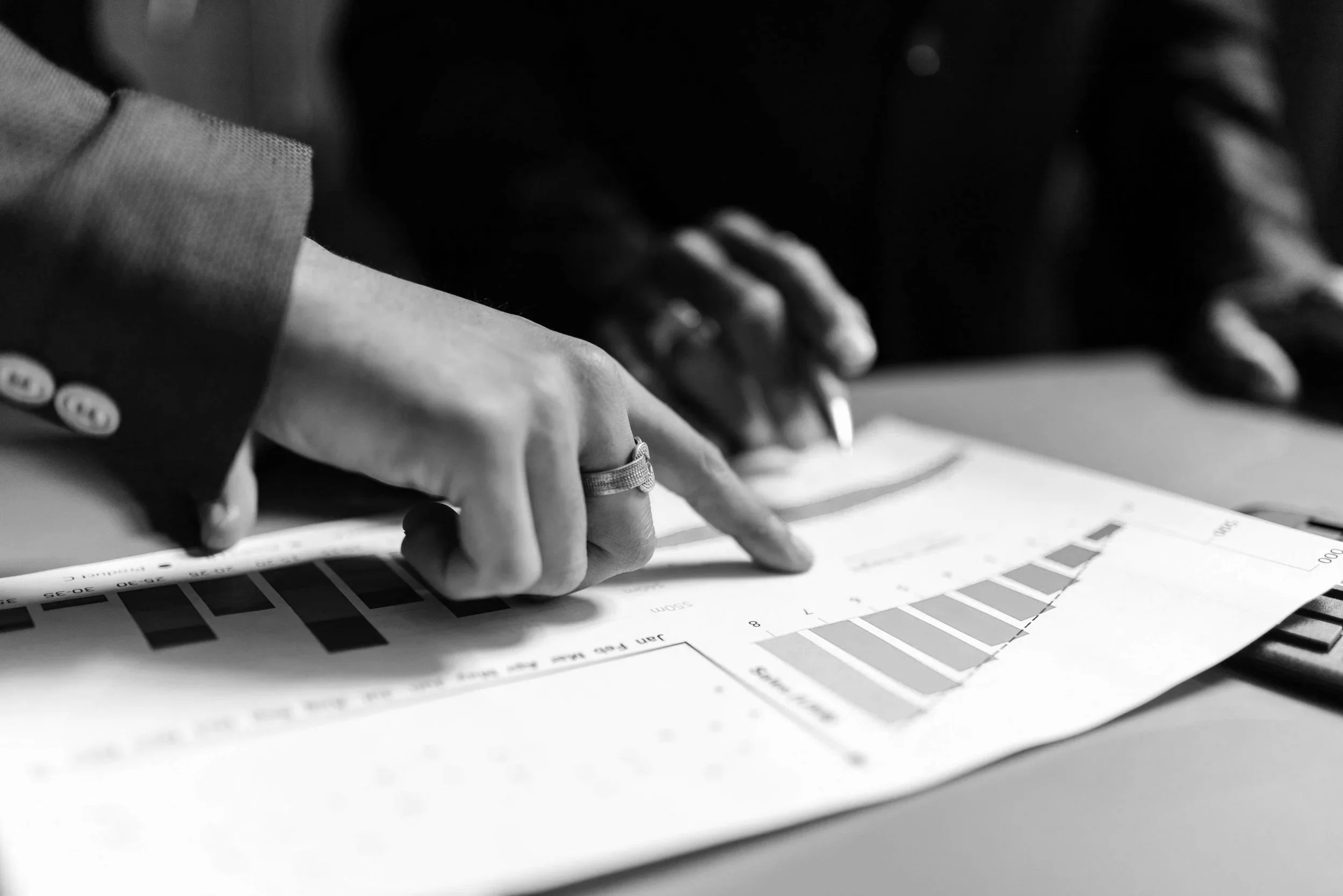 Close-up of two people pointing at a printed chart or graph on paper, with a focus on a person wearing a suit with a ring on their finger, in black and white.