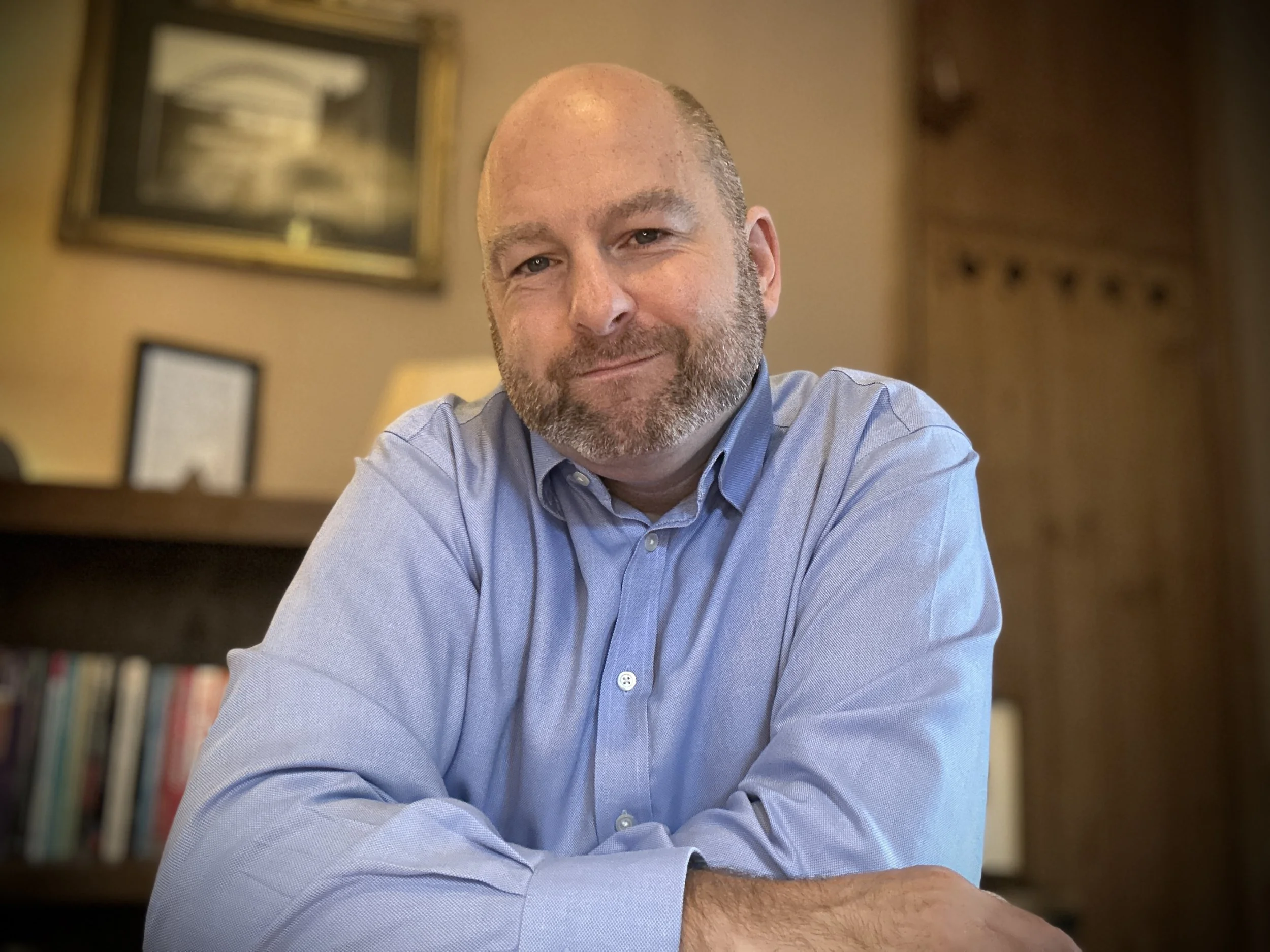 A bearded man in a light blue dress shirt smiling at the camera, sitting with arms crossed in a warmly lit room with wooden paneling and framed pictures or artwork in the background.