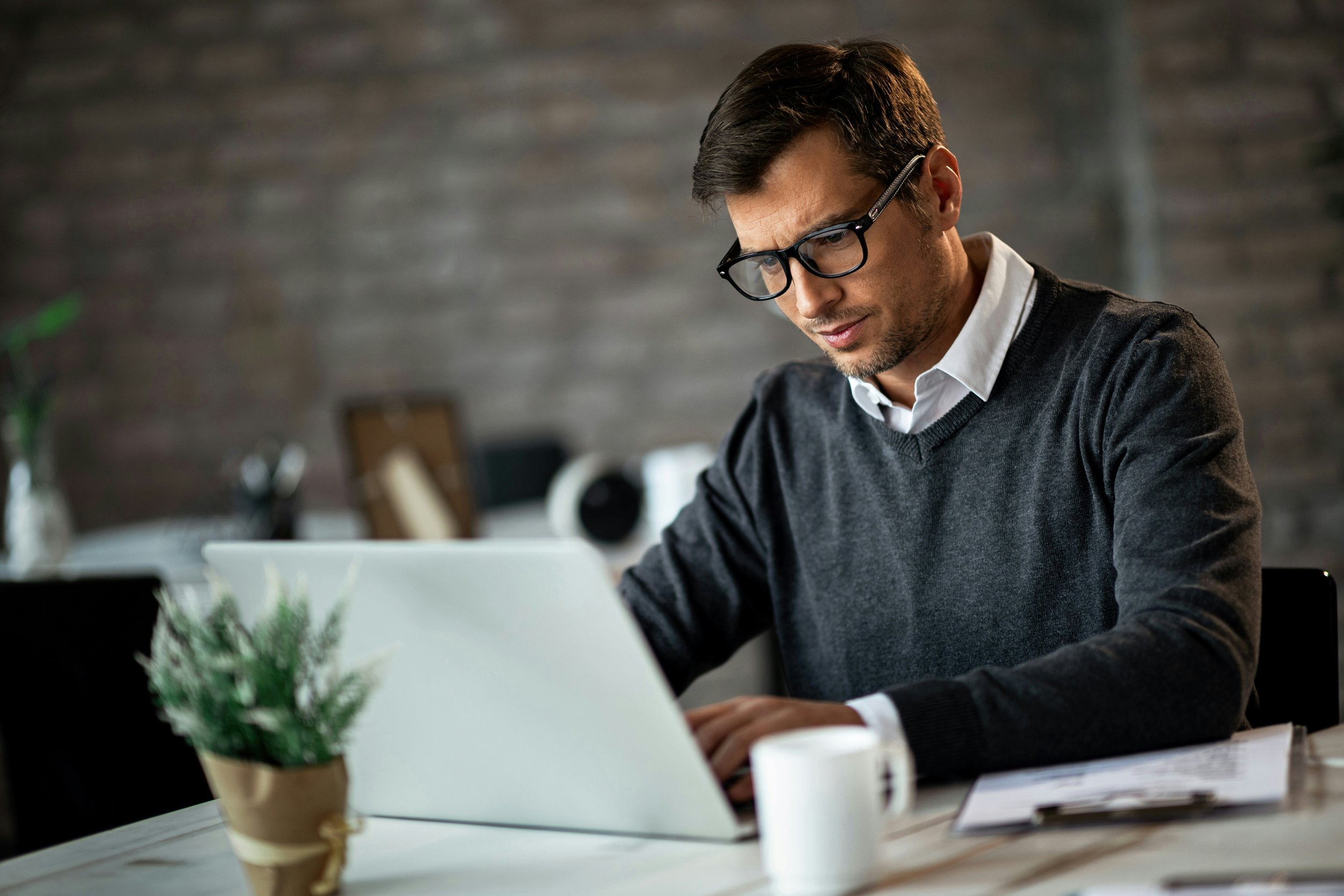 A man with glasses working at a desk in an office setting with a laptop, potted plant, and stacks of papers.