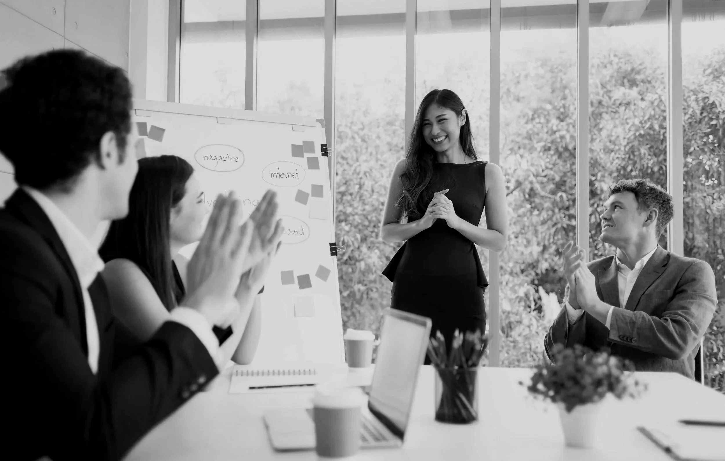 Black-and-white photo of a woman giving a presentation to a group of four people in a modern office with large windows. The woman is smiling and clapping, while the seated individuals are also clapping and smiling. There is a whiteboard with notes and sticky notes behind her, and various office items on the table, including laptops, notebooks, and cups.