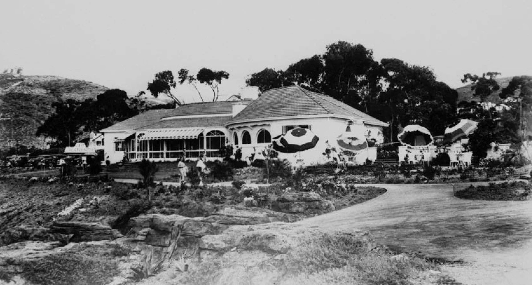 Black and white photo of the Victor Hugo Inn with a rounded, sloped roof and a large porch, surrounded by trees, umbrellas, and a garden, with people walking on a path in the foreground.