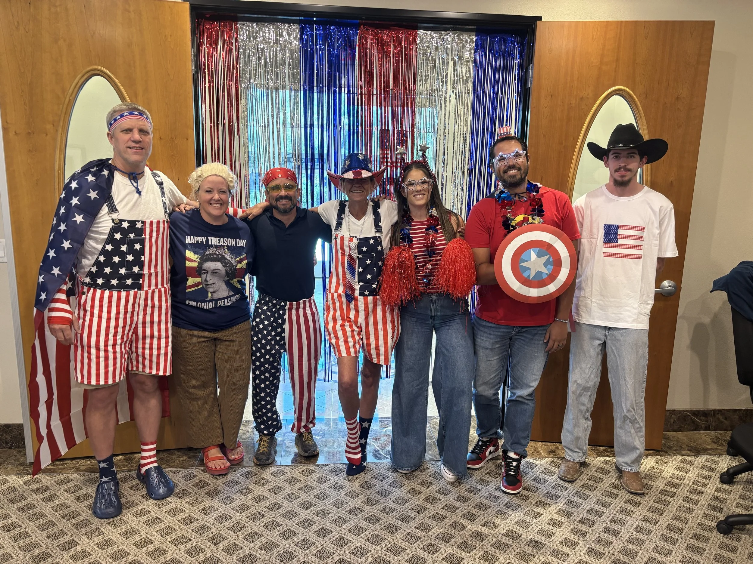 Group of seven people dressed in patriotic American-themed costumes standing in front of a shiny red, white, and blue curtain backdrop. They are smiling and appear to be celebrating.