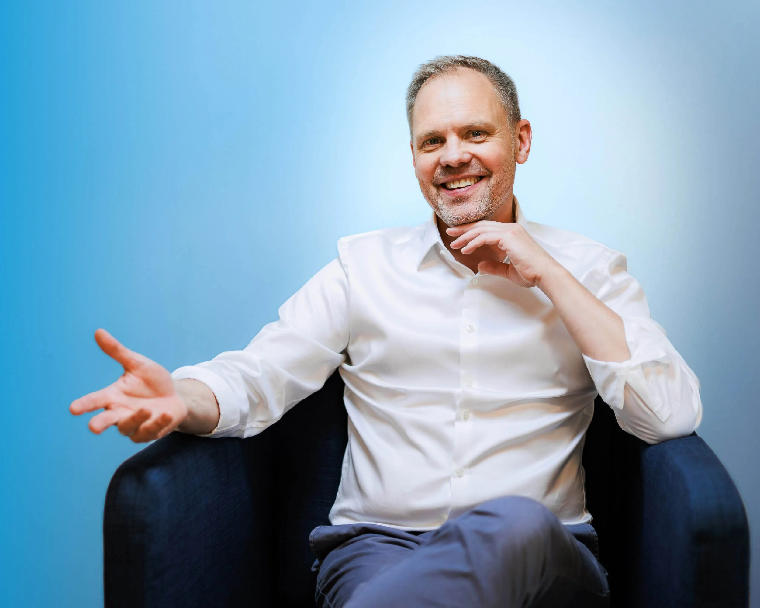 Businessfotograf Berlin: Thomas Dürst sitzend im Sessel mit offener Geste vor blauem Studiohintergrund