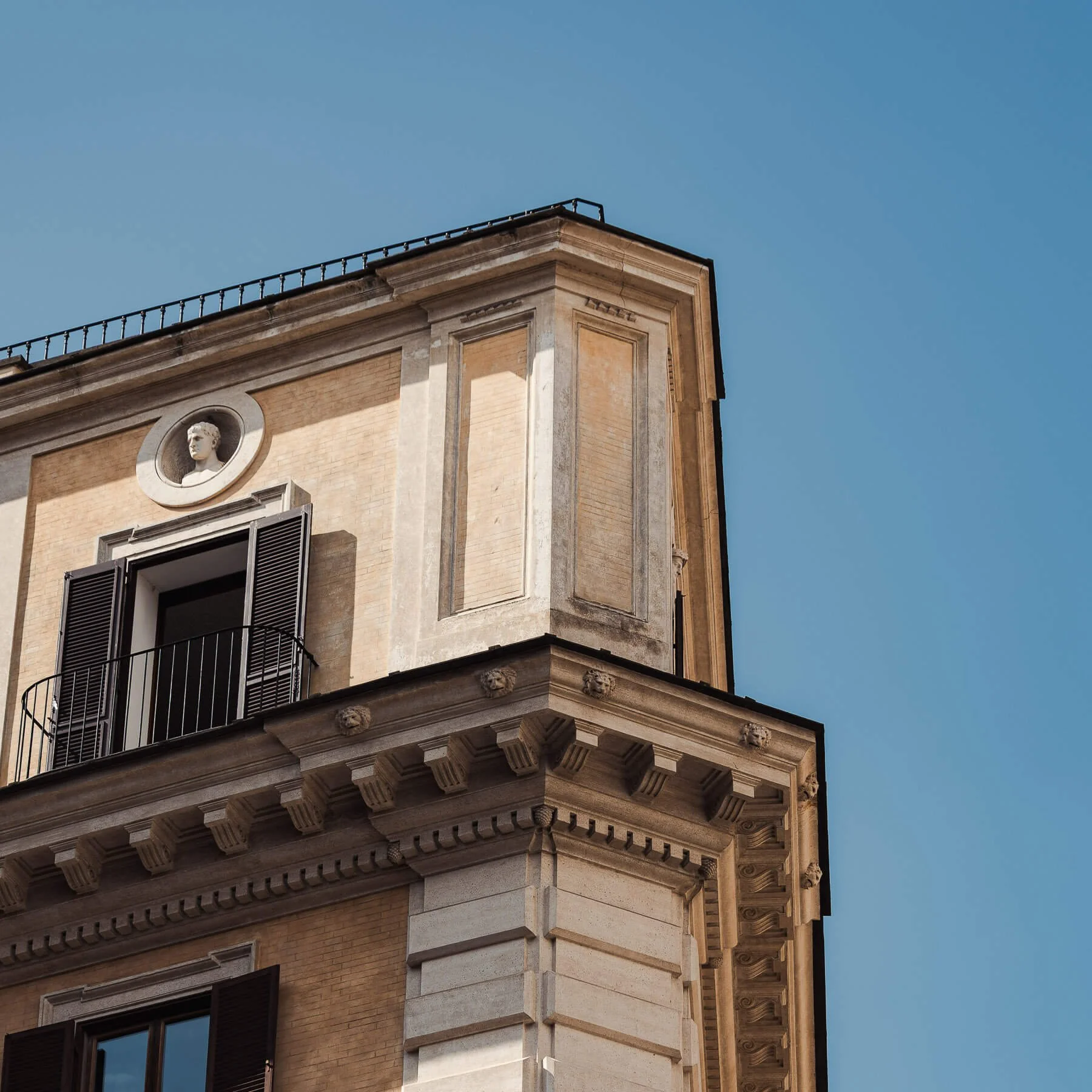 Architekturfotograf Berlin: Historische Gebäudeecke in Rom mit Balkon, Fensterläden und Büstenrelief unter blauem Himmel