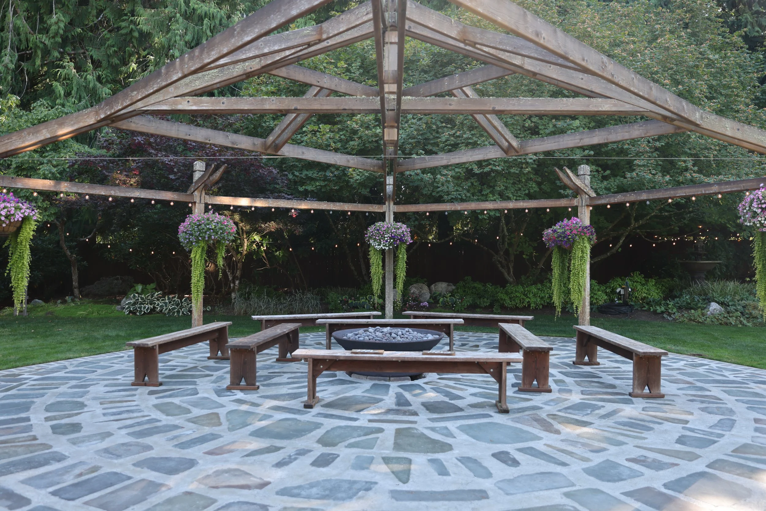Garden fire pit with flames and rocks, outdoor evening party scene with string lights, people socializing, wooden building, chairs, and trees in the background.
