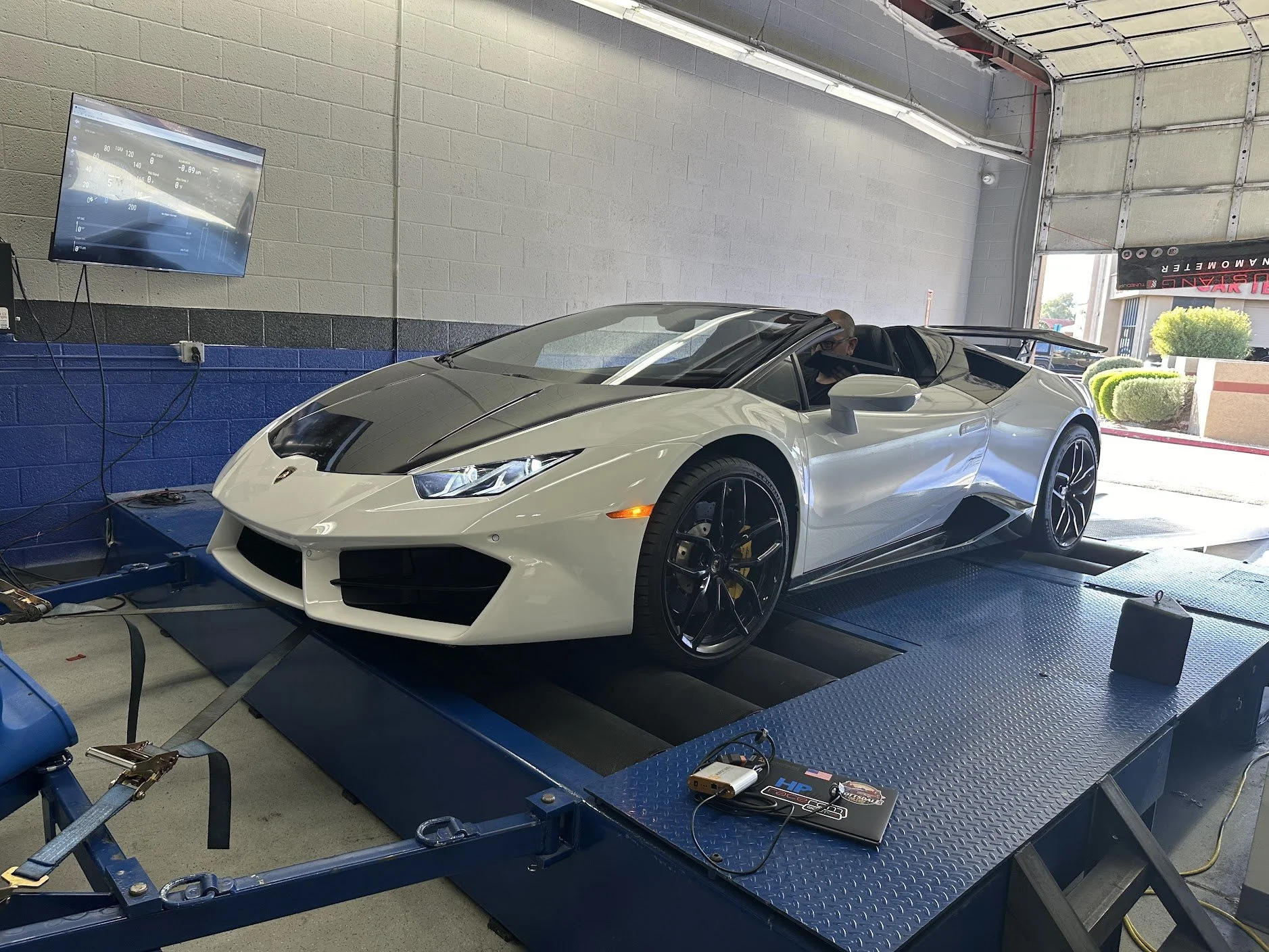 A white Lamborghini sports car with black racing stripes on a dyno testing platform indoors. A person is sitting in the driver’s seat. The room has a beige and blue wall, a flat-screen monitor, and various electronic equipment on the platform.