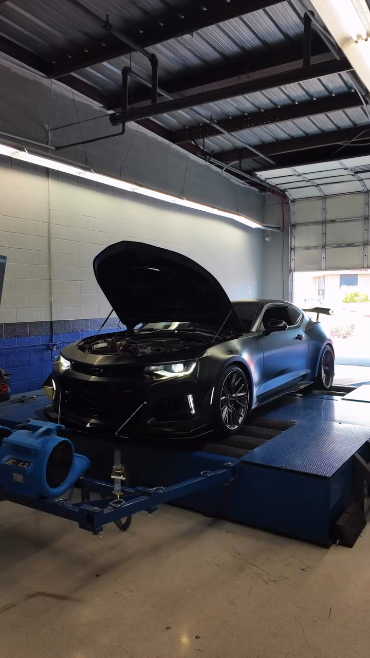 A black sports car with its hood open, positioned on a dynamometer in a garage or industrial setting.