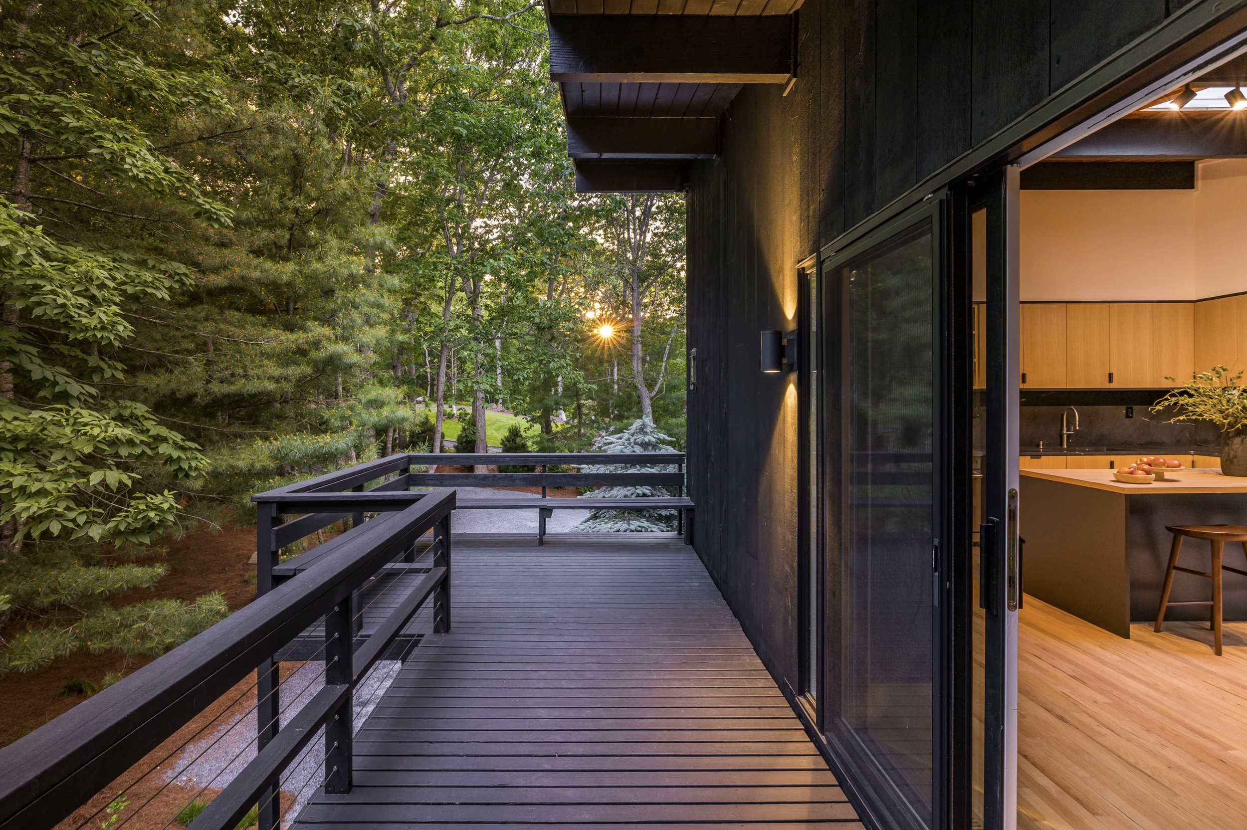 Covered exterior walkway with dark-stained wood siding and railing, looking into a softly lit kitchen through large sliding glass doors, surrounded by mature trees.
