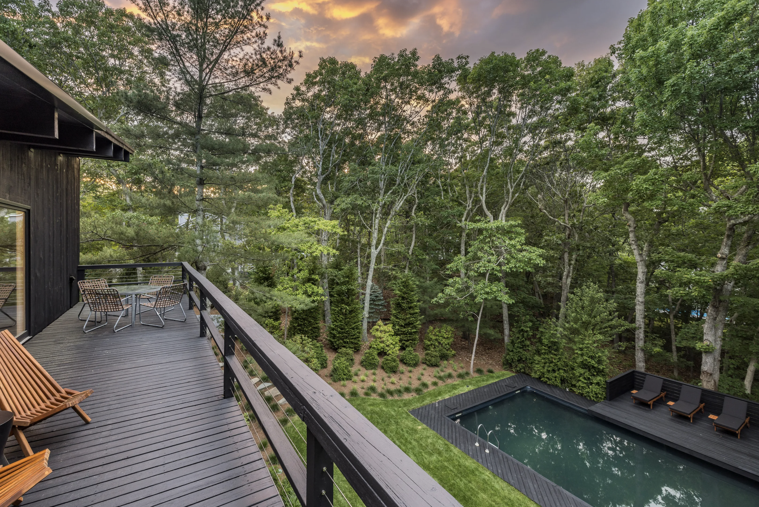 Upper-level wood deck overlooking a rectangular pool and forested yard, furnished with lounge chairs and outdoor seating at sunset