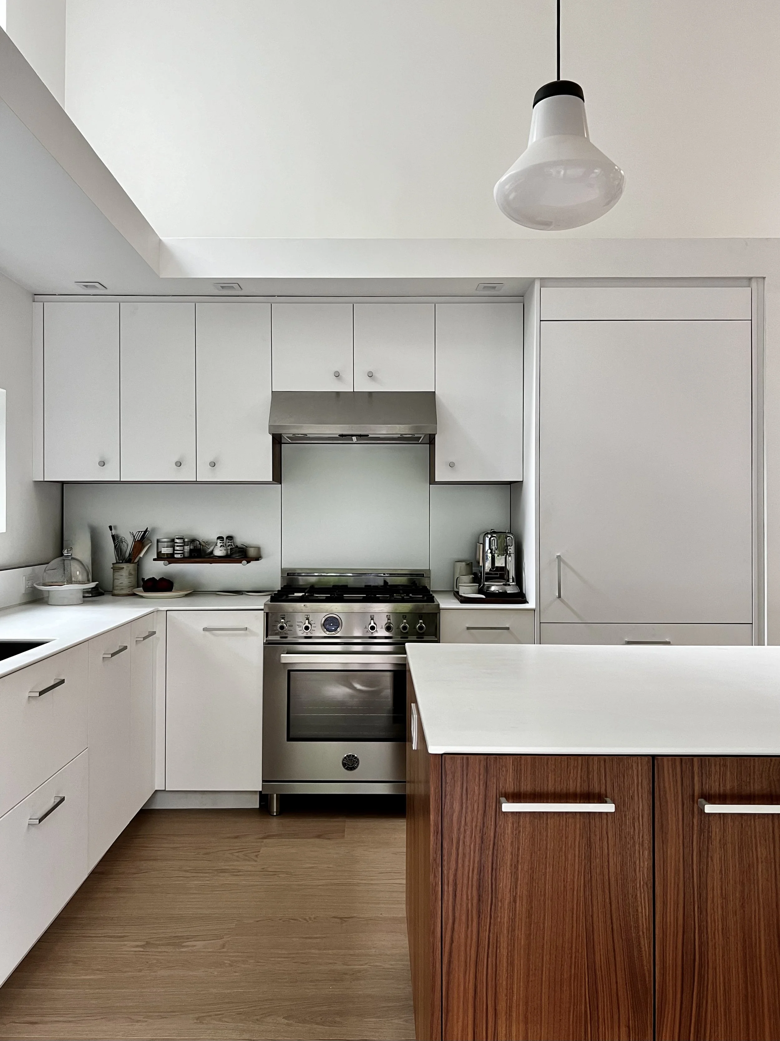 Minimalist kitchen with white flat-panel cabinetry, stainless steel range, pale glass backsplash, and a warm wood island topped with a light stone surface, illuminated by a simple pendant light.