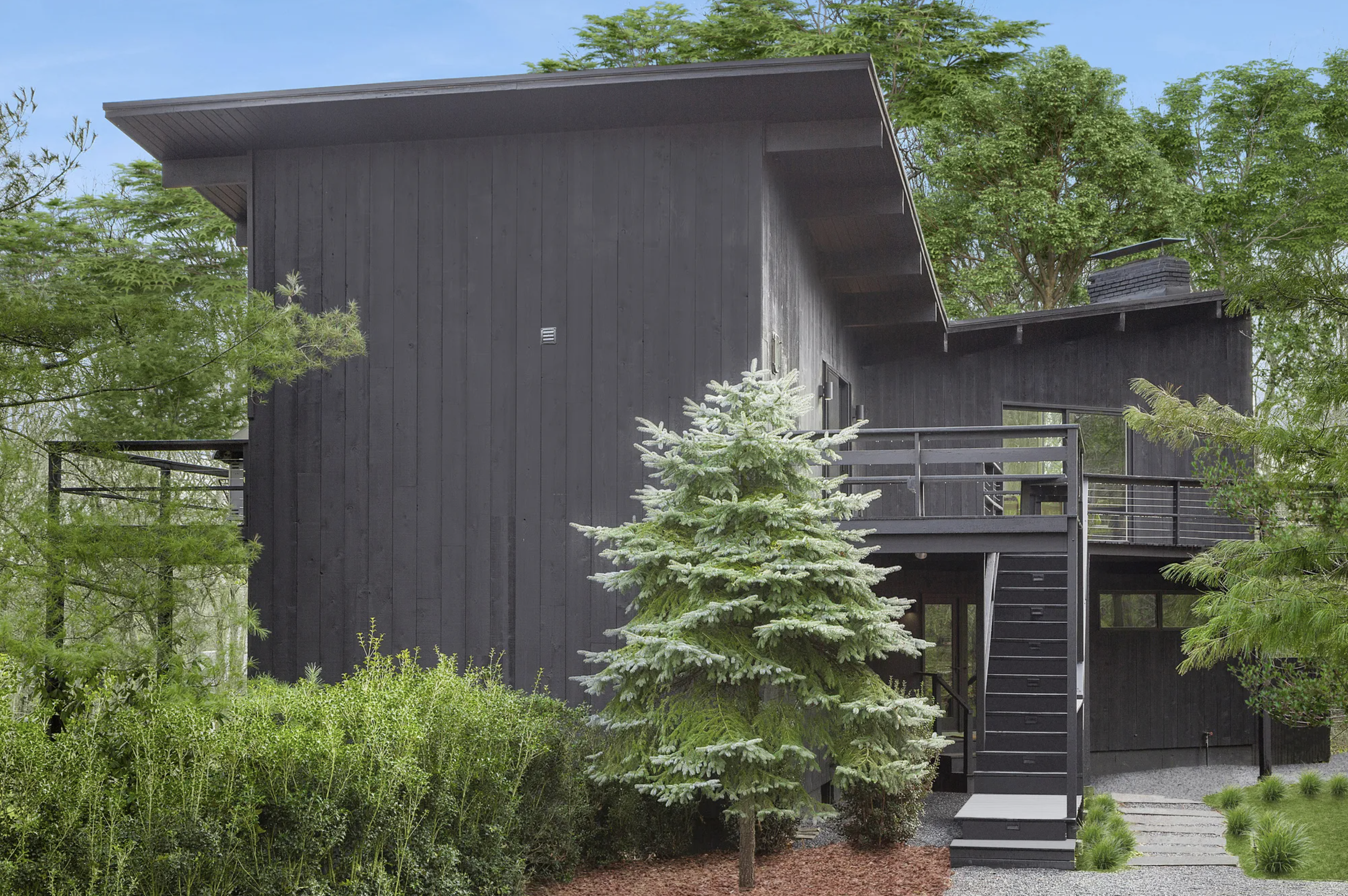 Modern, dark-clad two-story home set among trees, featuring a raised deck, exterior staircase, and landscaped woodland setting