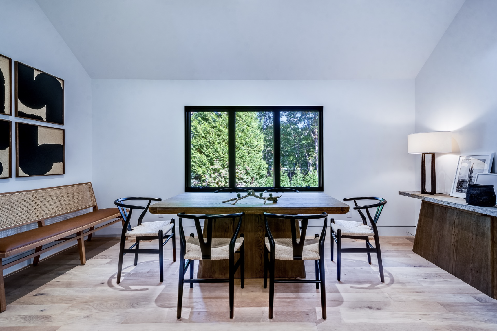 Minimalist dining room with solid wood table, black dining chairs, light wood floors, and large picture window framing lush greenery