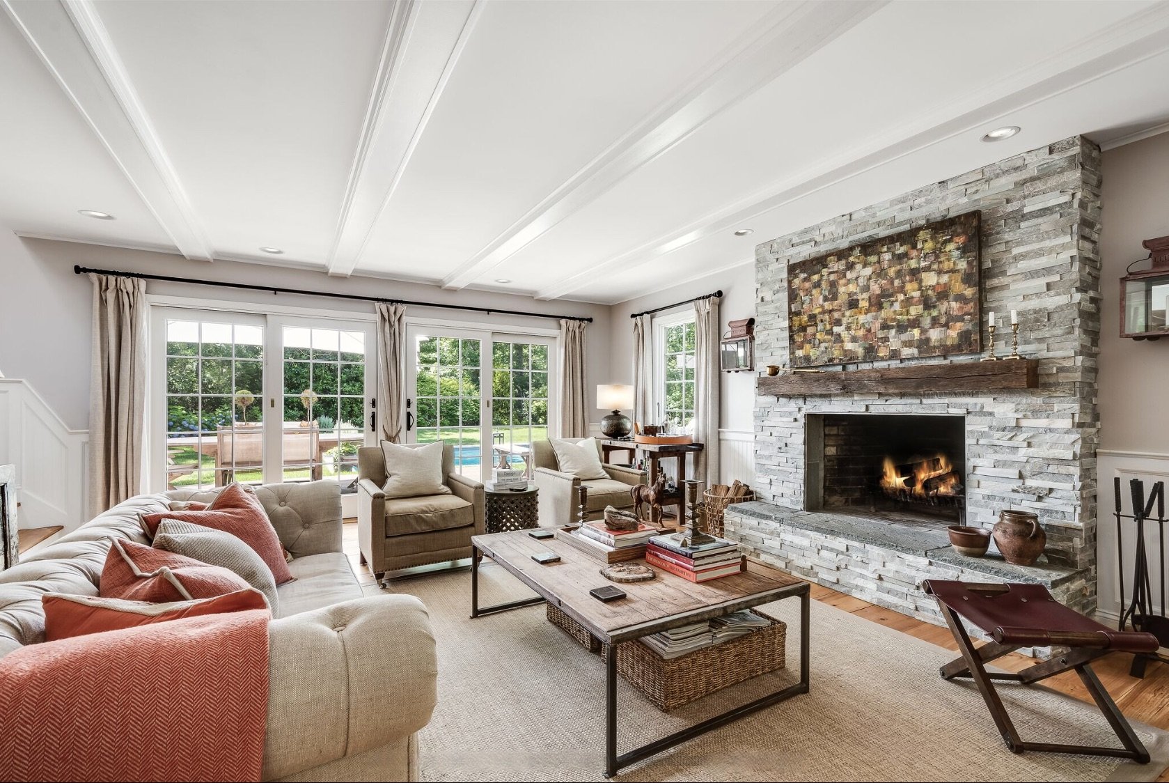 A warm, light-filled living room with exposed ceiling beams, a stone fireplace with a wood mantel, neutral upholstered seating, and French doors opening to a landscaped backyard and pool